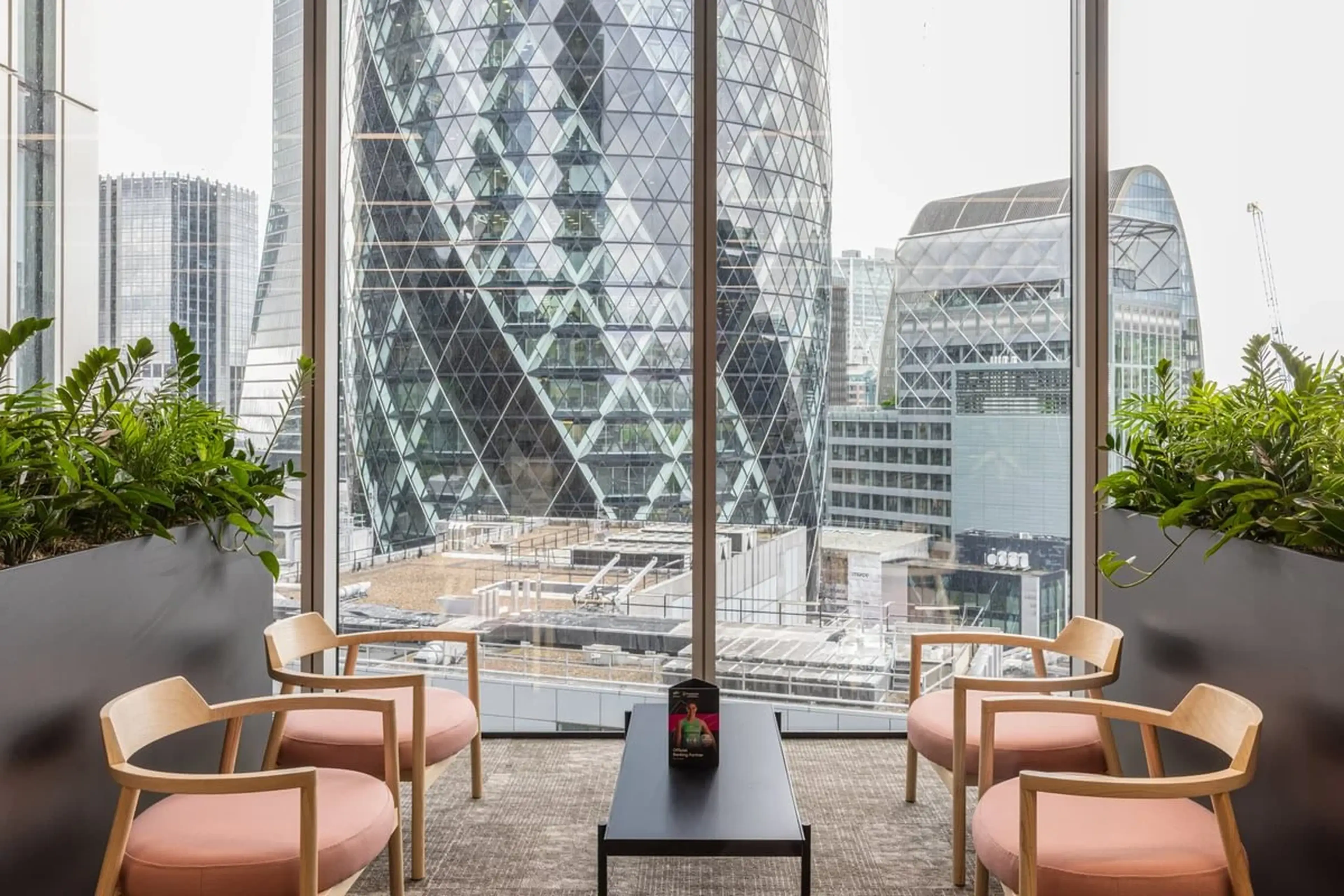 Modern office lounge with four chairs and a table, overlooking city skyscrapers through large windows, surrounded by indoor plants.