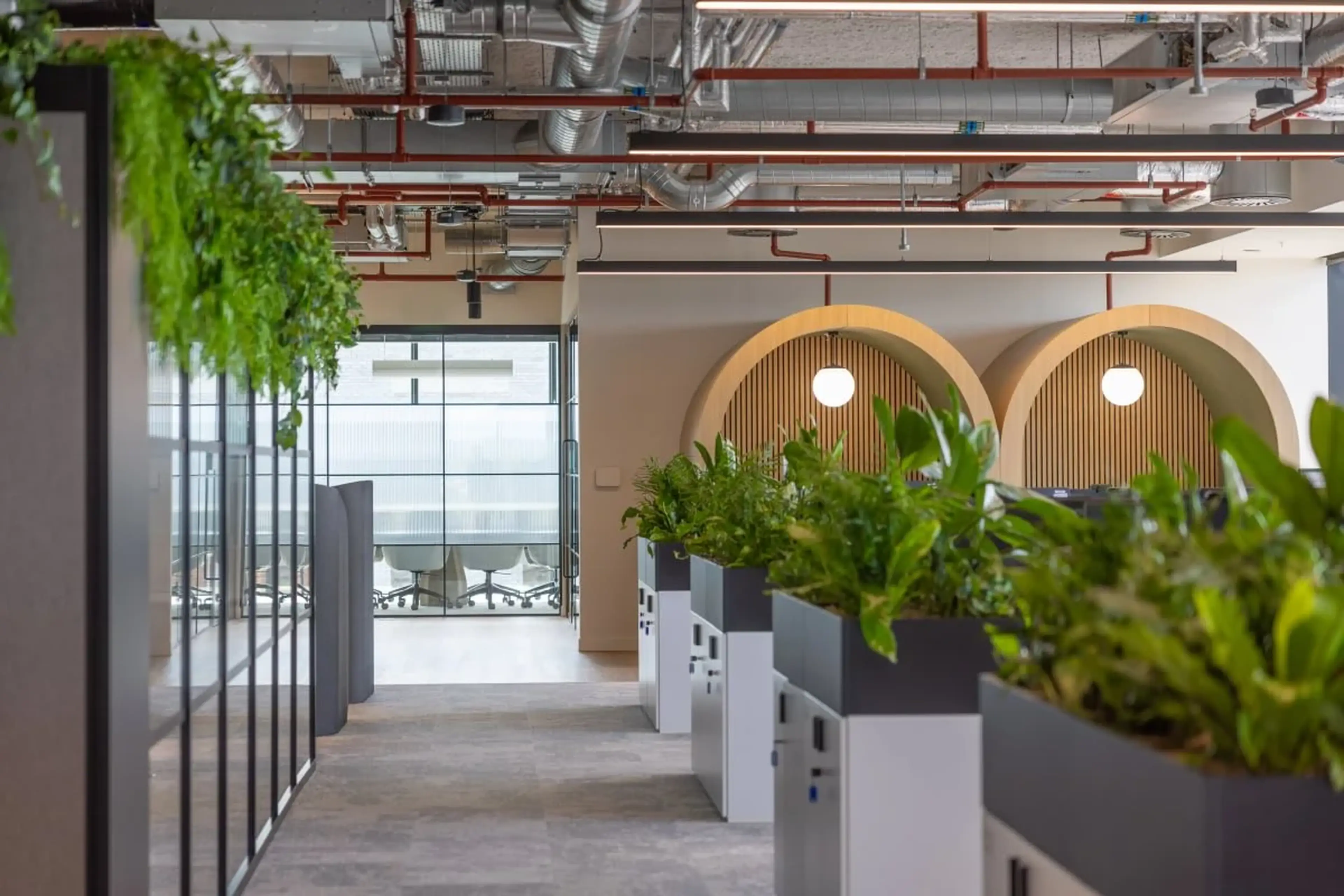 Modern office space with indoor plants, glass partitions, wooden arch decor, and visible ceiling pipes, leading to a conference room.