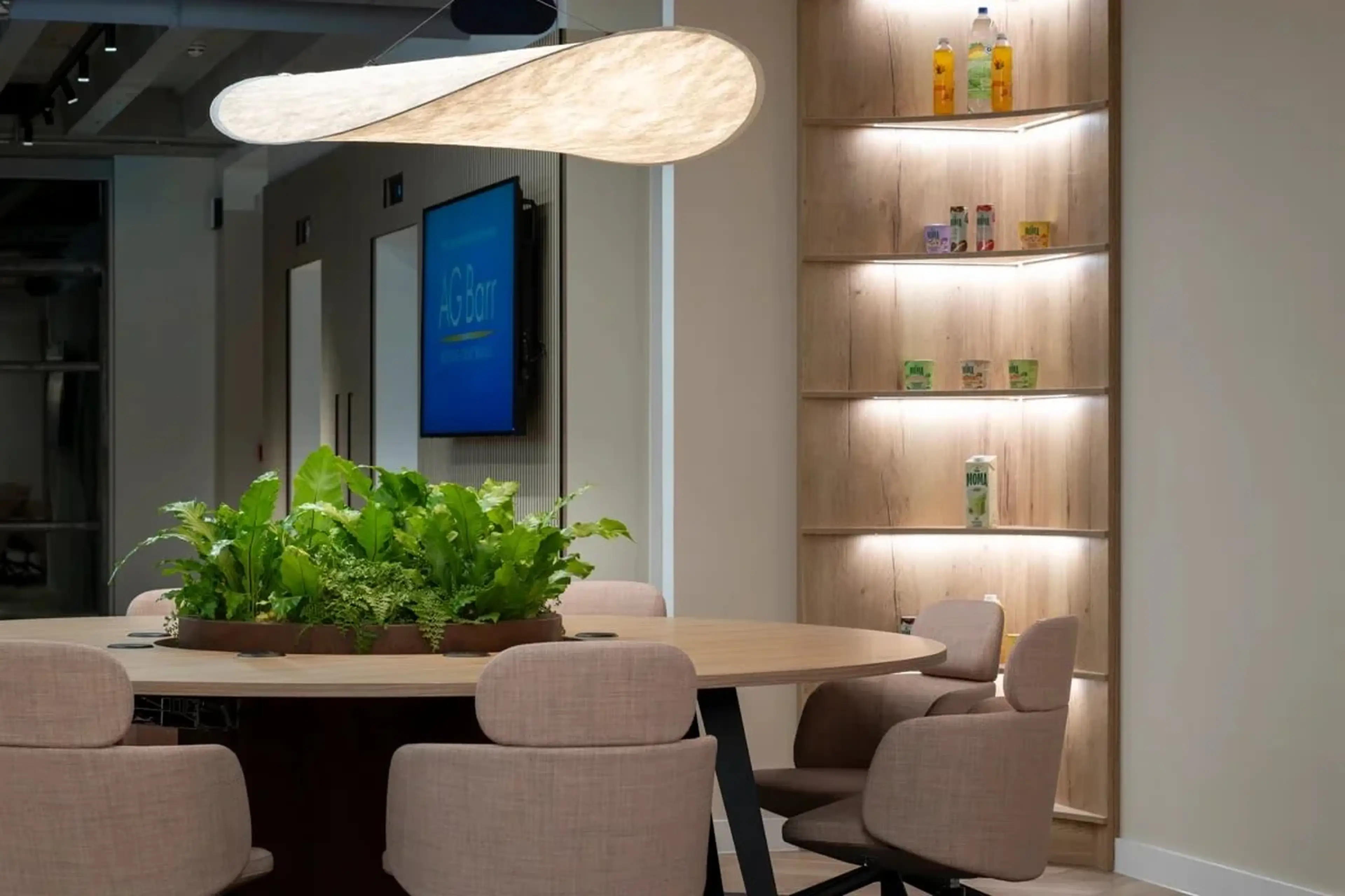 Modern office space with a round table and plants, surrounded by beige chairs. A unique overhead light fixture and lit shelves with decor and bottles.