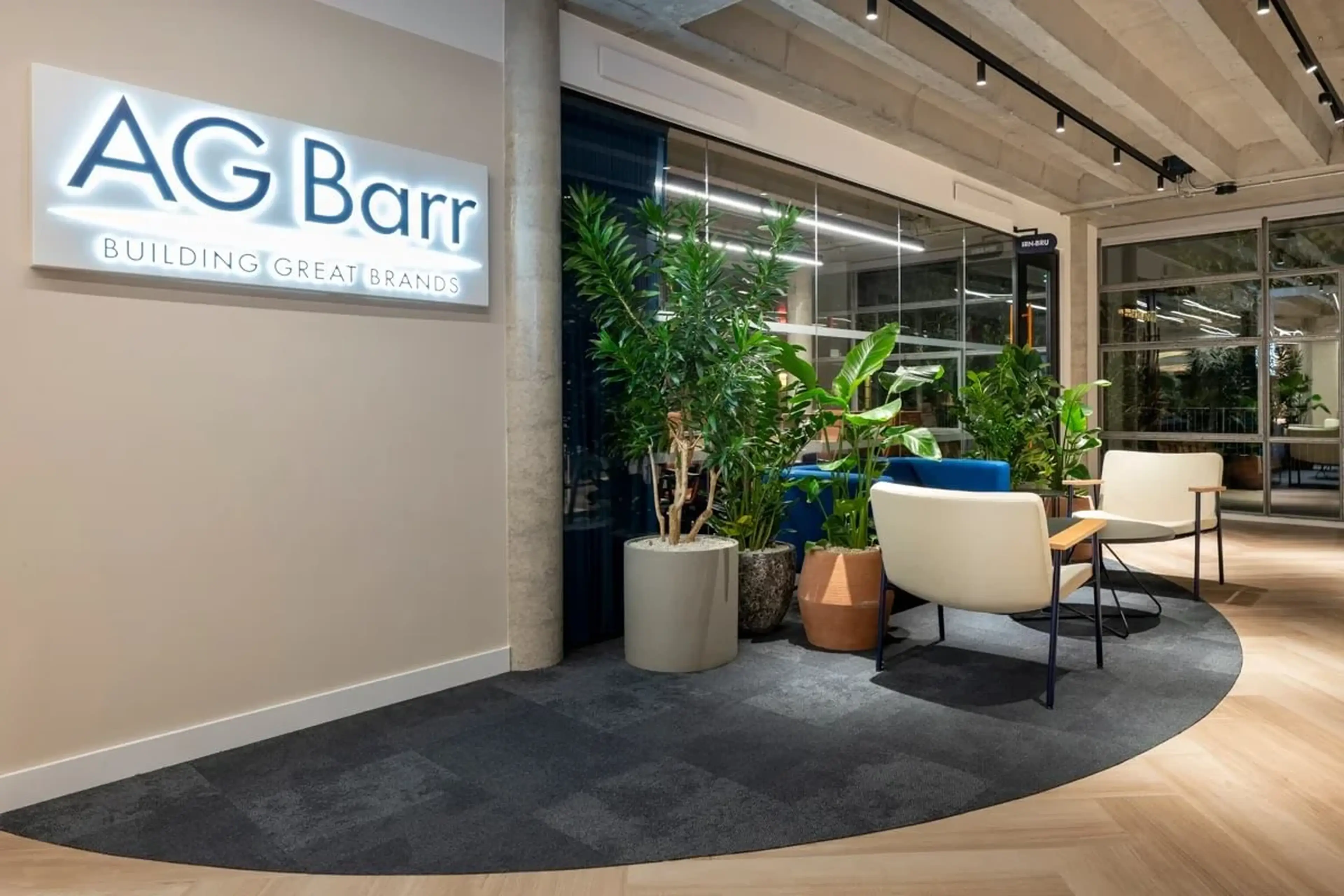 Modern office reception area with "AG Barr" sign, chairs, tables, and lush indoor plants on a dark carpeted section of the wood floor.