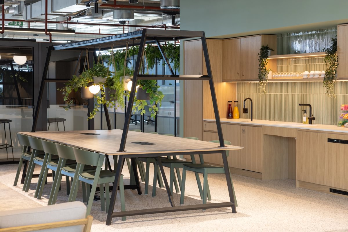 Modern office kitchen with a large table, hanging plants, and a light fixture. Wooden cabinets and countertops with sinks in the background.