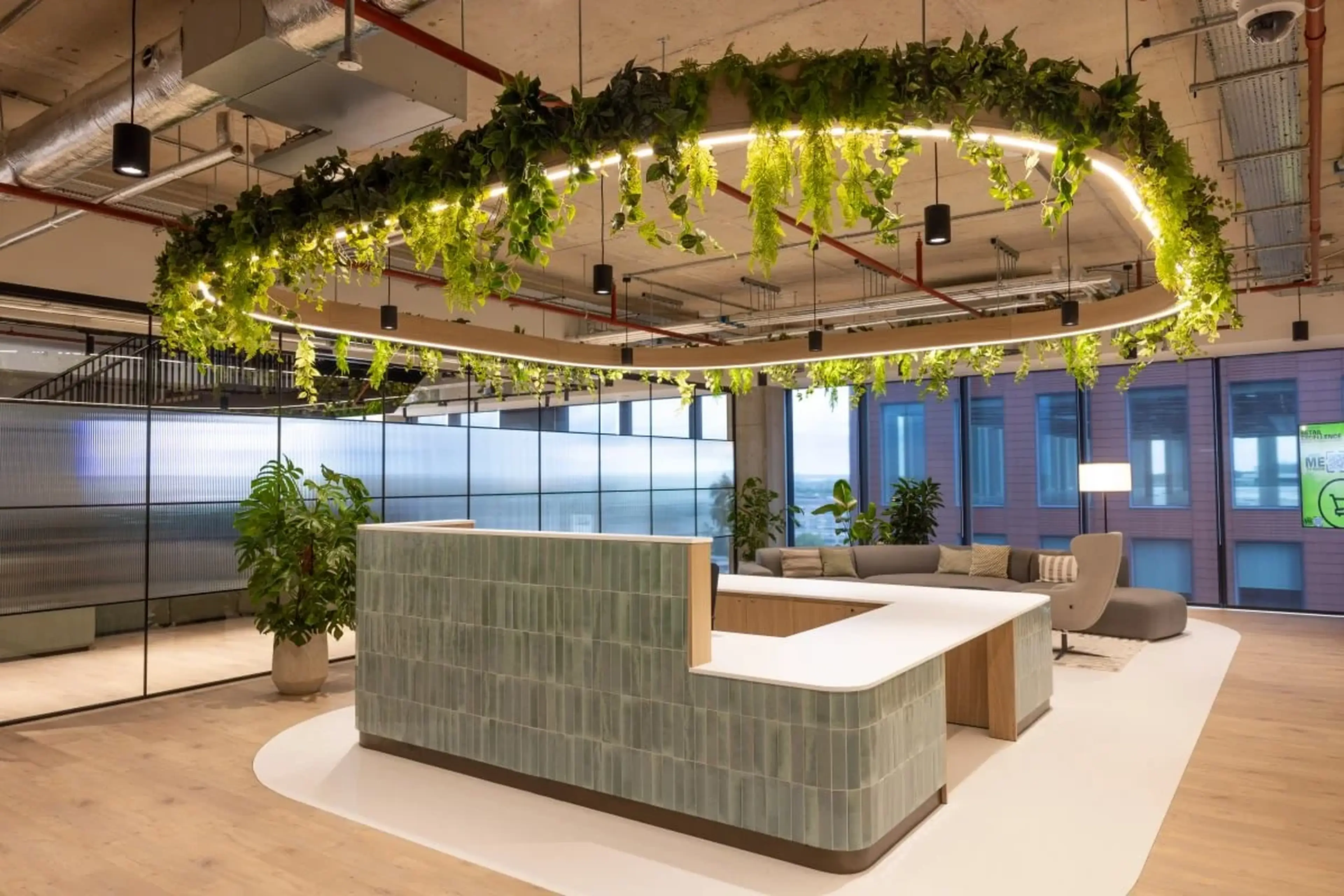 Modern office lobby with a curved reception desk, hanging greenery, and plants. Glass walls and ceiling lights create a bright, open atmosphere.