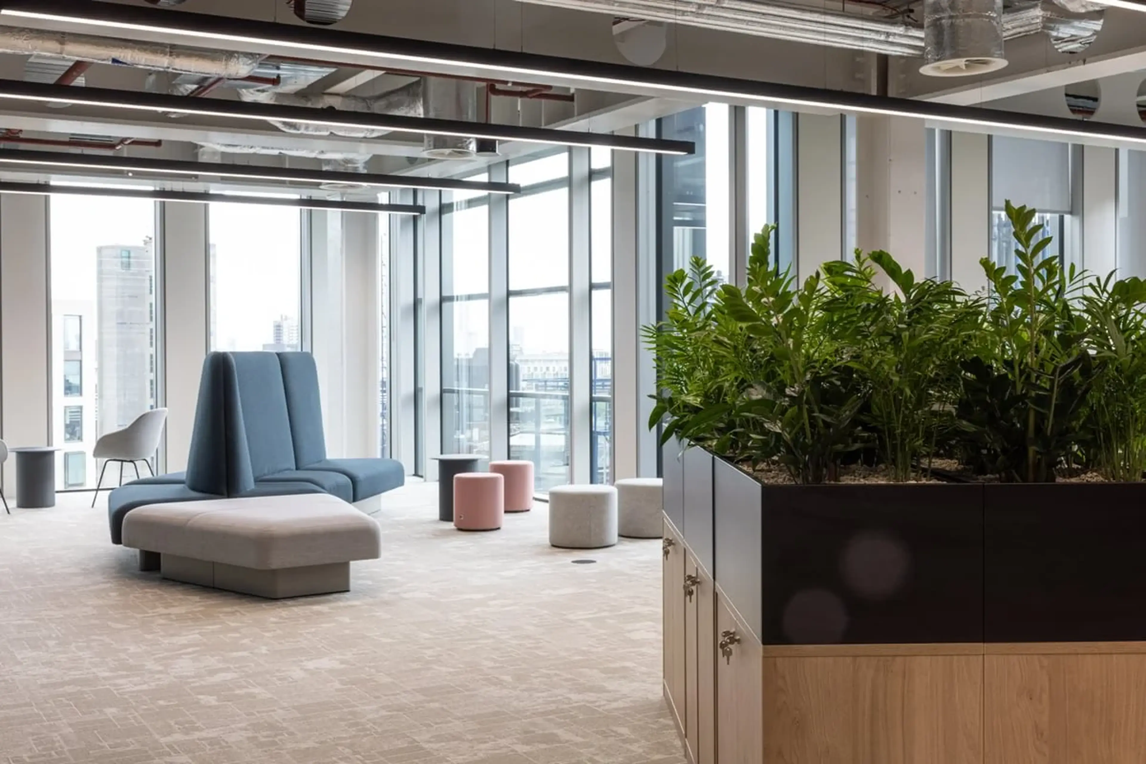 Modern office lounge with large windows, blue and gray seating, pink stools, and a planter with lush green plants.