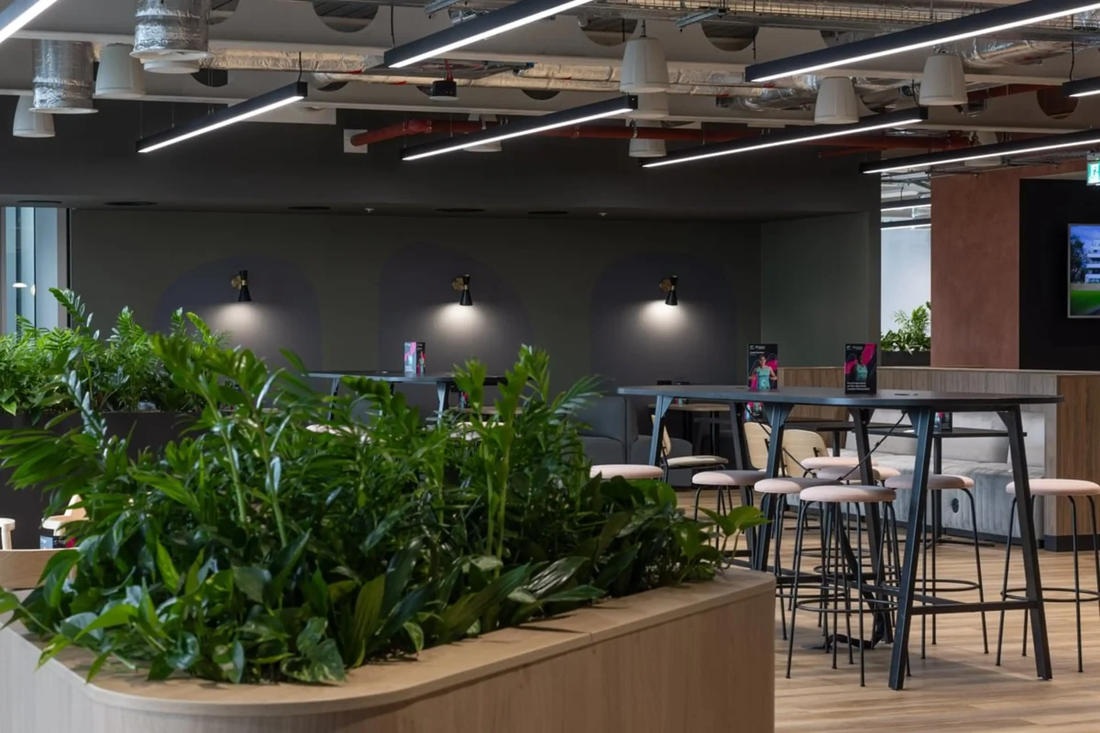 Modern office space with high tables, stools, and lush green plants. Overhead lighting and a TV screen in the background.