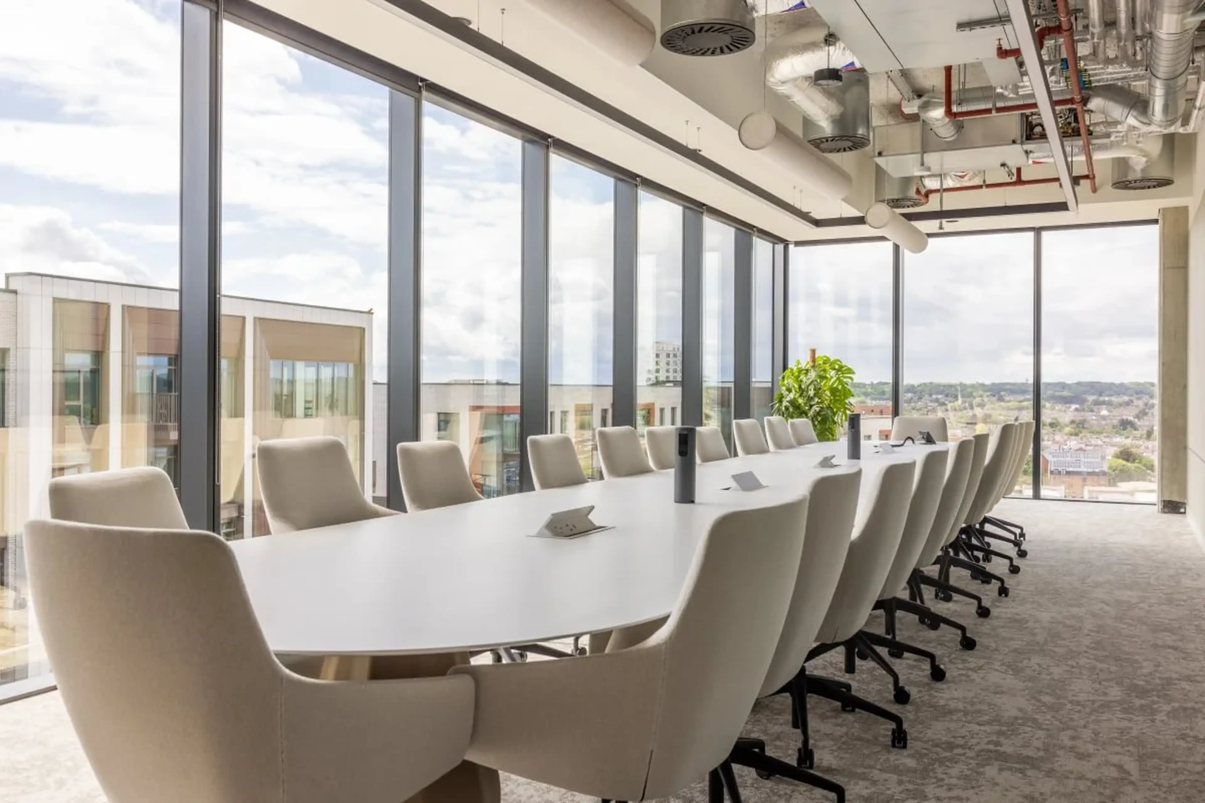 Modern conference room with a long white table, beige chairs, large windows, and city view. Exposed ceiling ducts add an industrial touch.