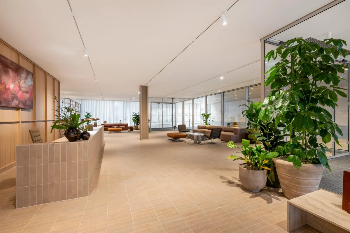 Spacious modern lobby with sofas, plants, glass walls, and a reception desk. Neutral tones and contemporary artwork enhance the elegant design.