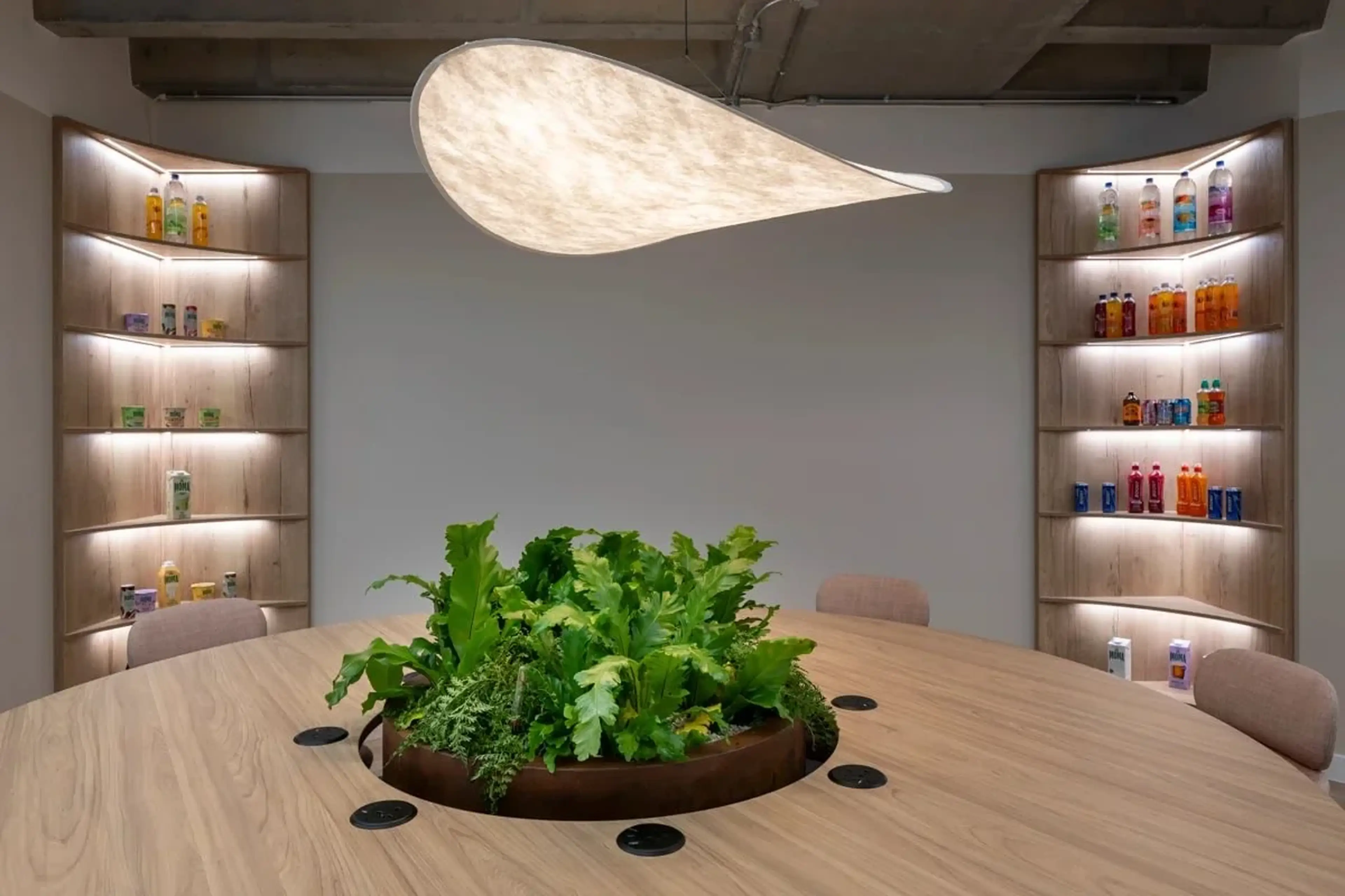 Modern meeting room with a round table featuring a plant centerpiece. Curved shelves display colorful bottles. Unique teardrop ceiling light above.
