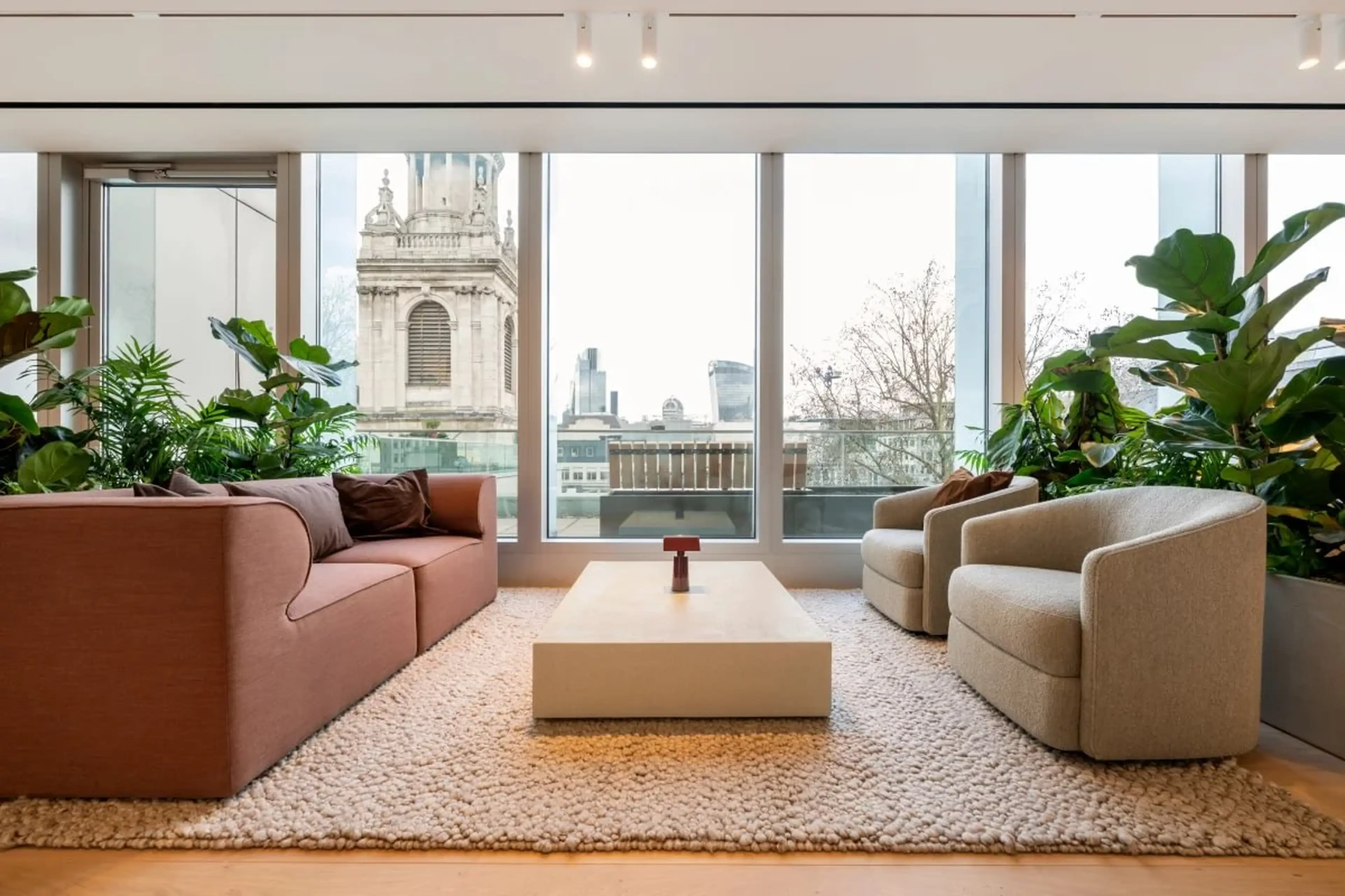 Modern living room with large windows, a pink sofa, beige chairs, a coffee table, plants, and a cityscape view featuring a historic building.