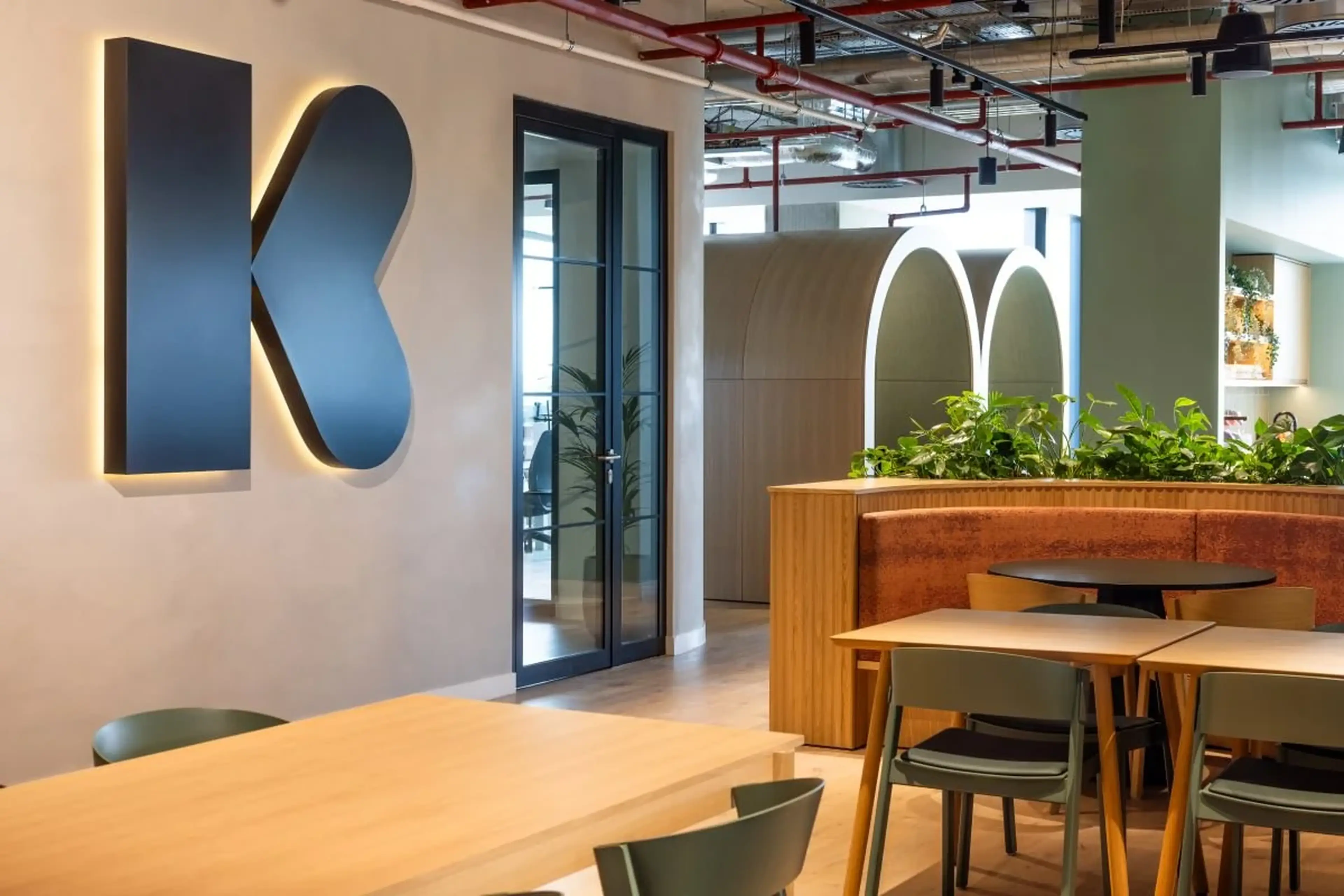 Modern office space with a large "K" on the wall, wooden tables, chairs, and greenery, featuring a glass door and arched structures.