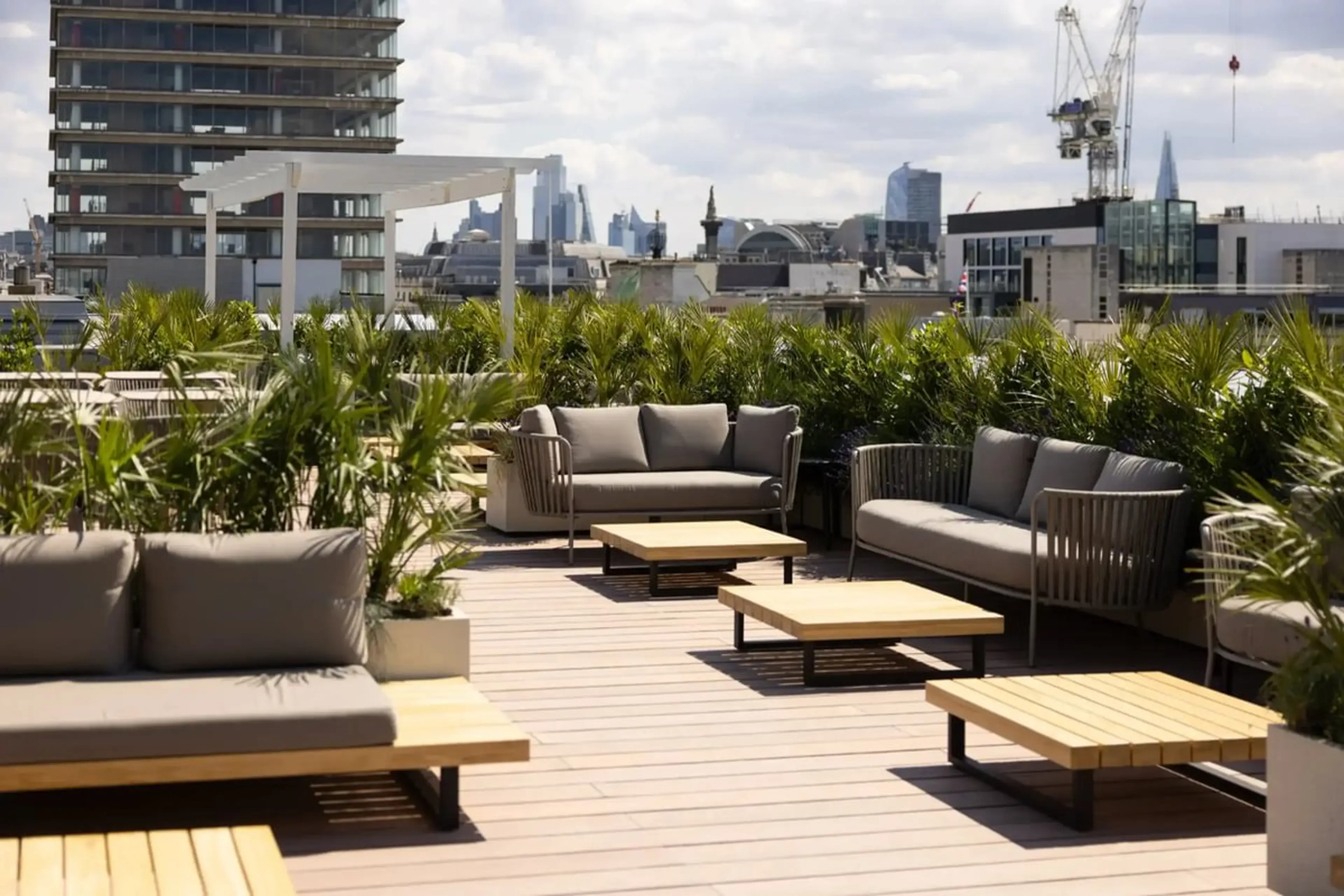 Rooftop patio with modern sofas, coffee tables, and surrounding greenery, overlooking city skyline on a partly cloudy day.