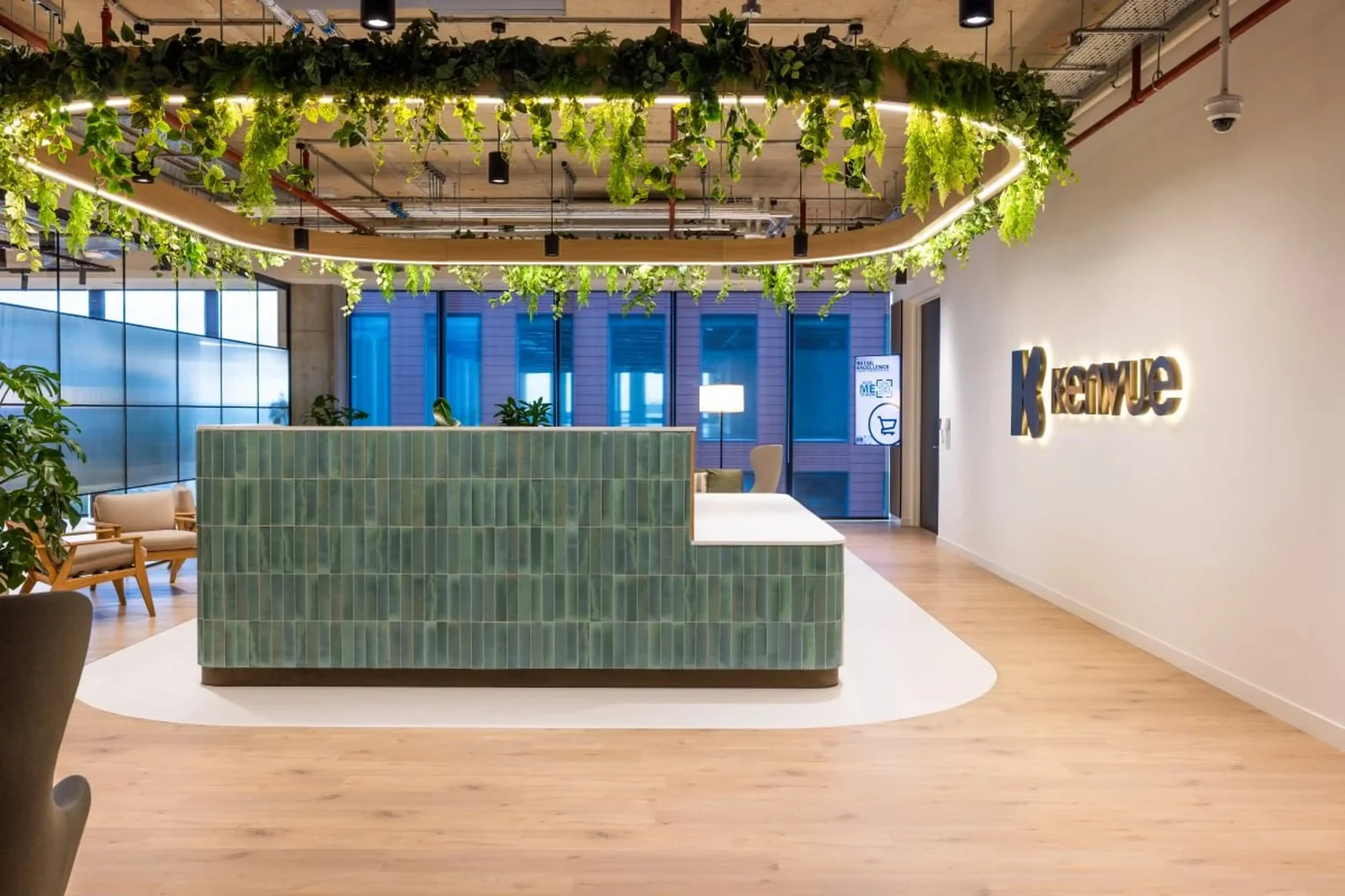 Modern office lobby with green tiled reception desk, overhead plants, and wooden flooring, featuring "Kenvue" logo on the wall.