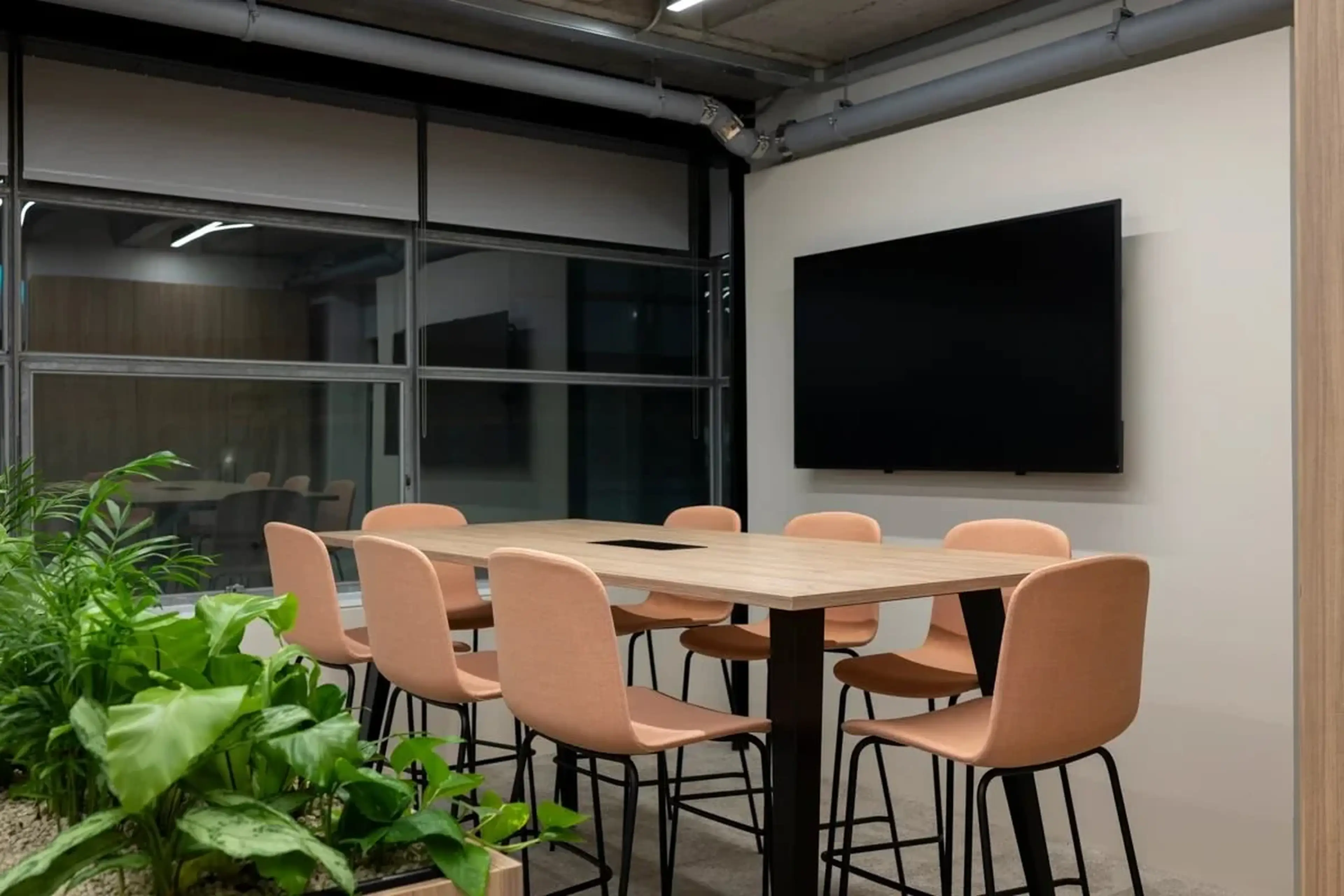 Modern conference room with a wooden table, eight peach chairs, a large screen on the wall, and indoor plants by the window.