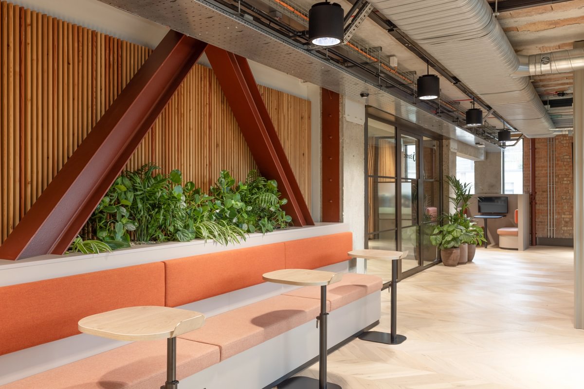 Modern office lounge with orange seating, small round tables, and indoor plants. Exposed ductwork and wood paneling enhance the contemporary design.