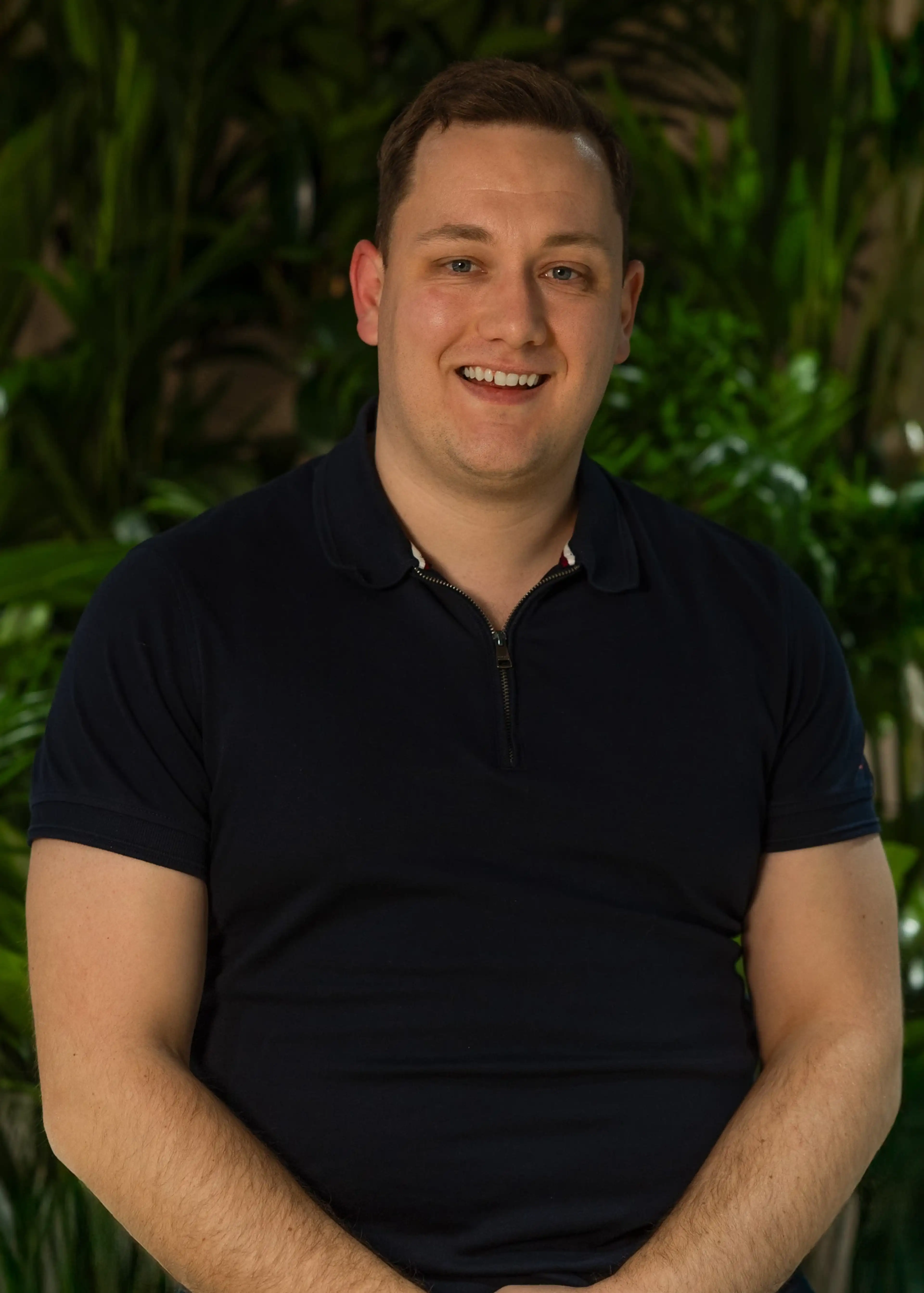 Smiling person in a navy shirt stands against a background of green foliage.
