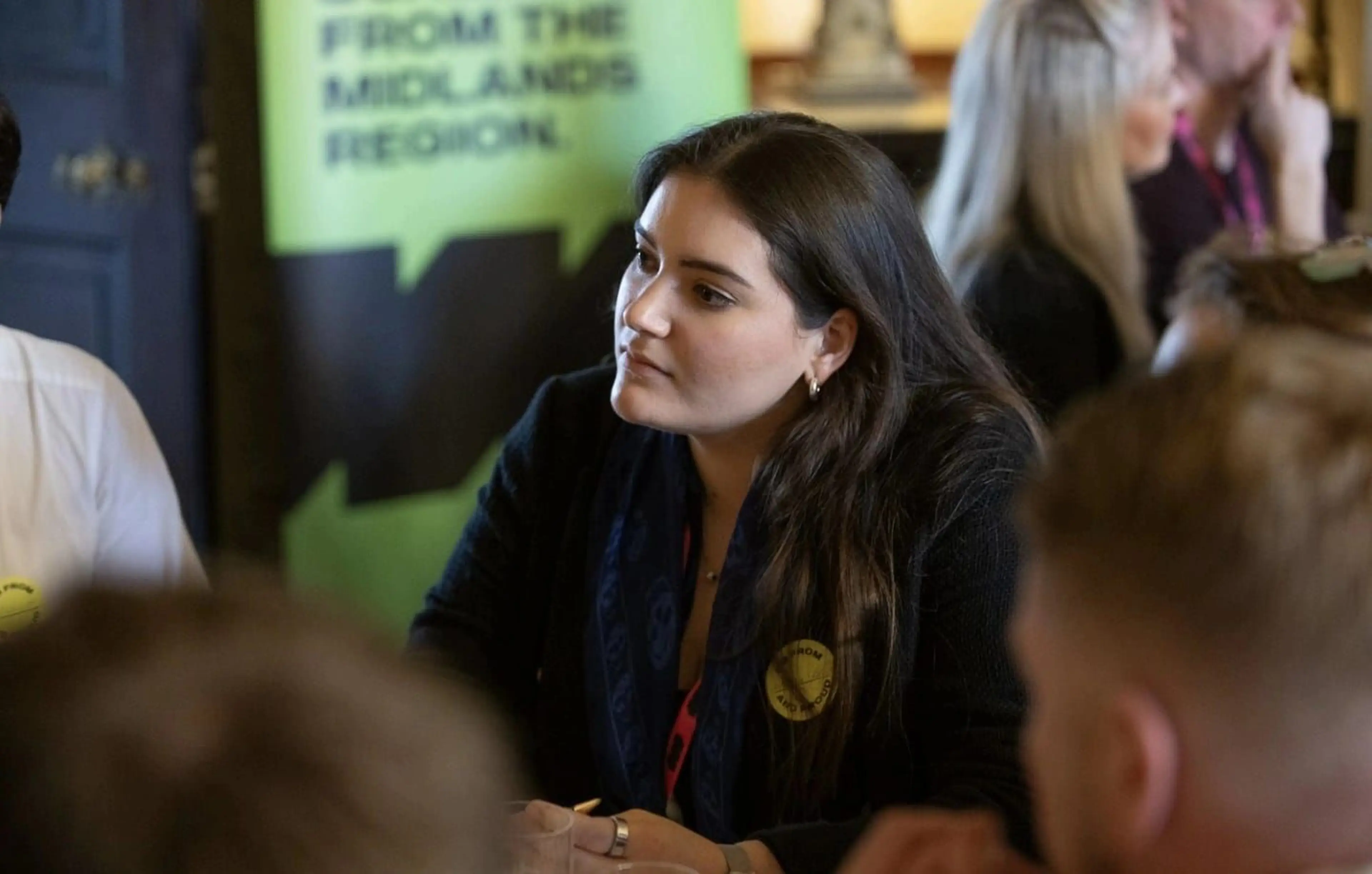 A woman in discussion, listening intently at a meeting. A green banner in the background reads, "From the Midlands Region."