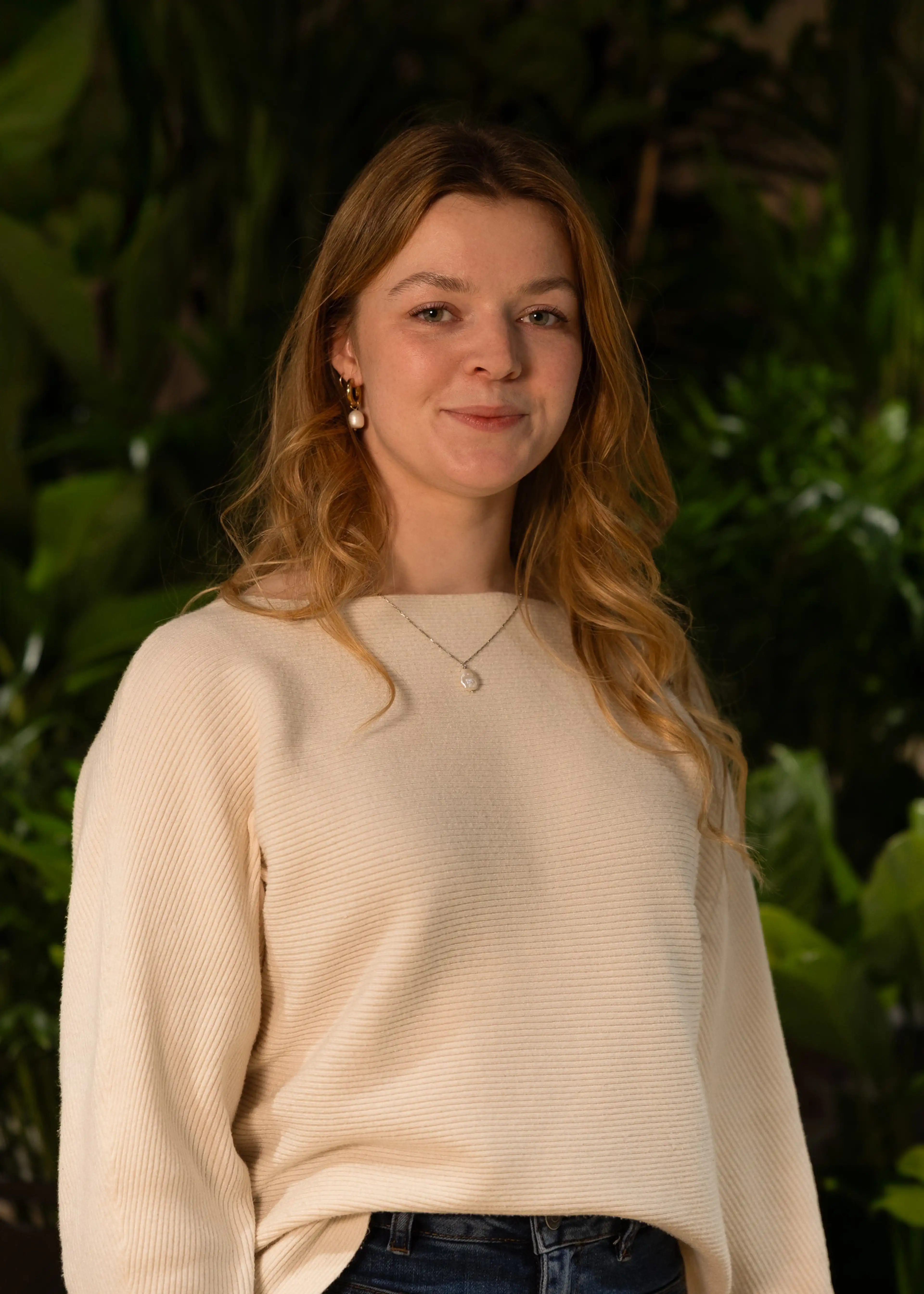 A woman with long blonde hair, wearing a cream sweater and jeans, stands smiling in front of lush green plants.
