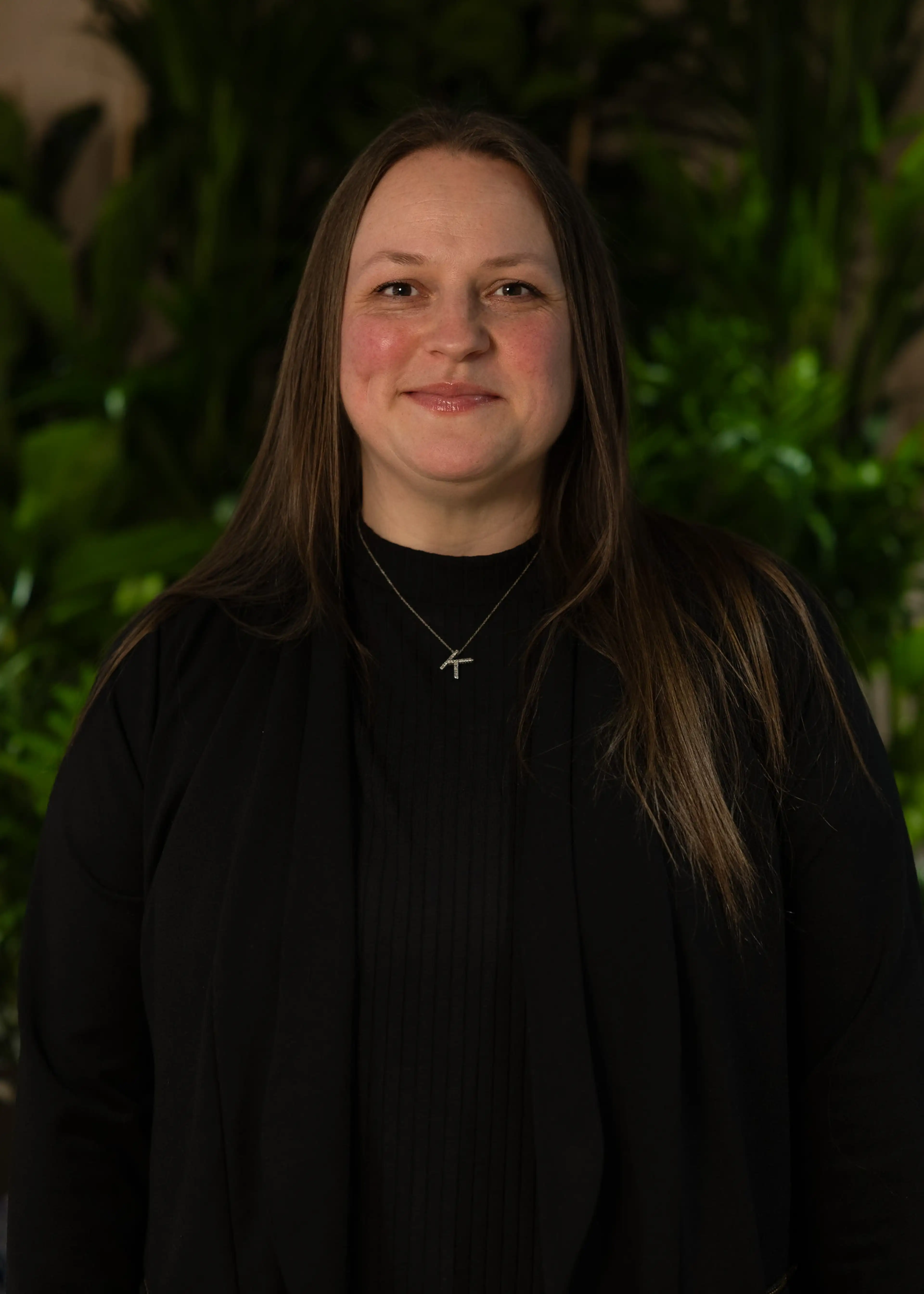 A person with long brown hair wearing a black outfit and a necklace, standing in front of lush green plants, smiling slightly.