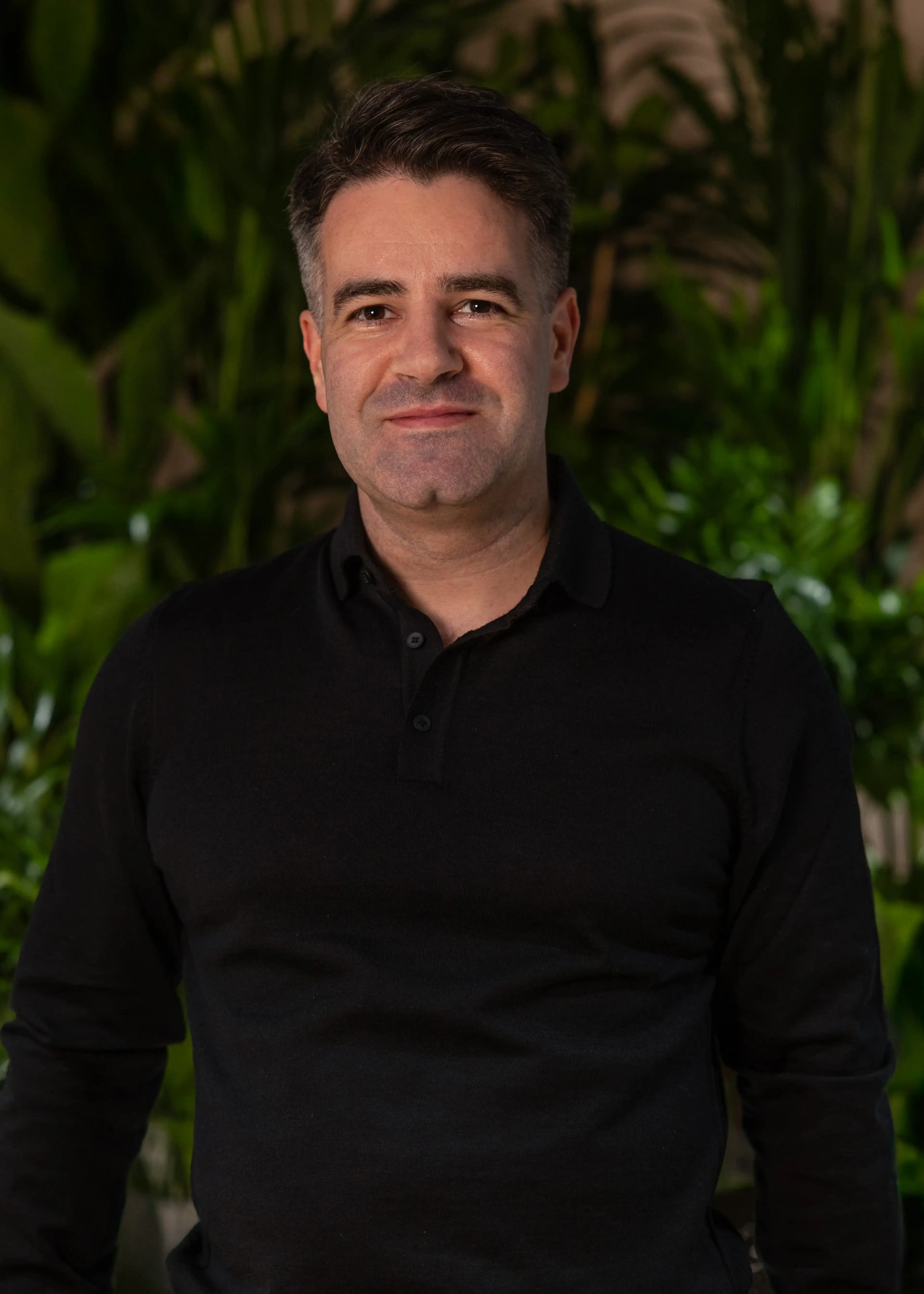 A person in a black long-sleeve shirt stands smiling, with green foliage in the blurred background.