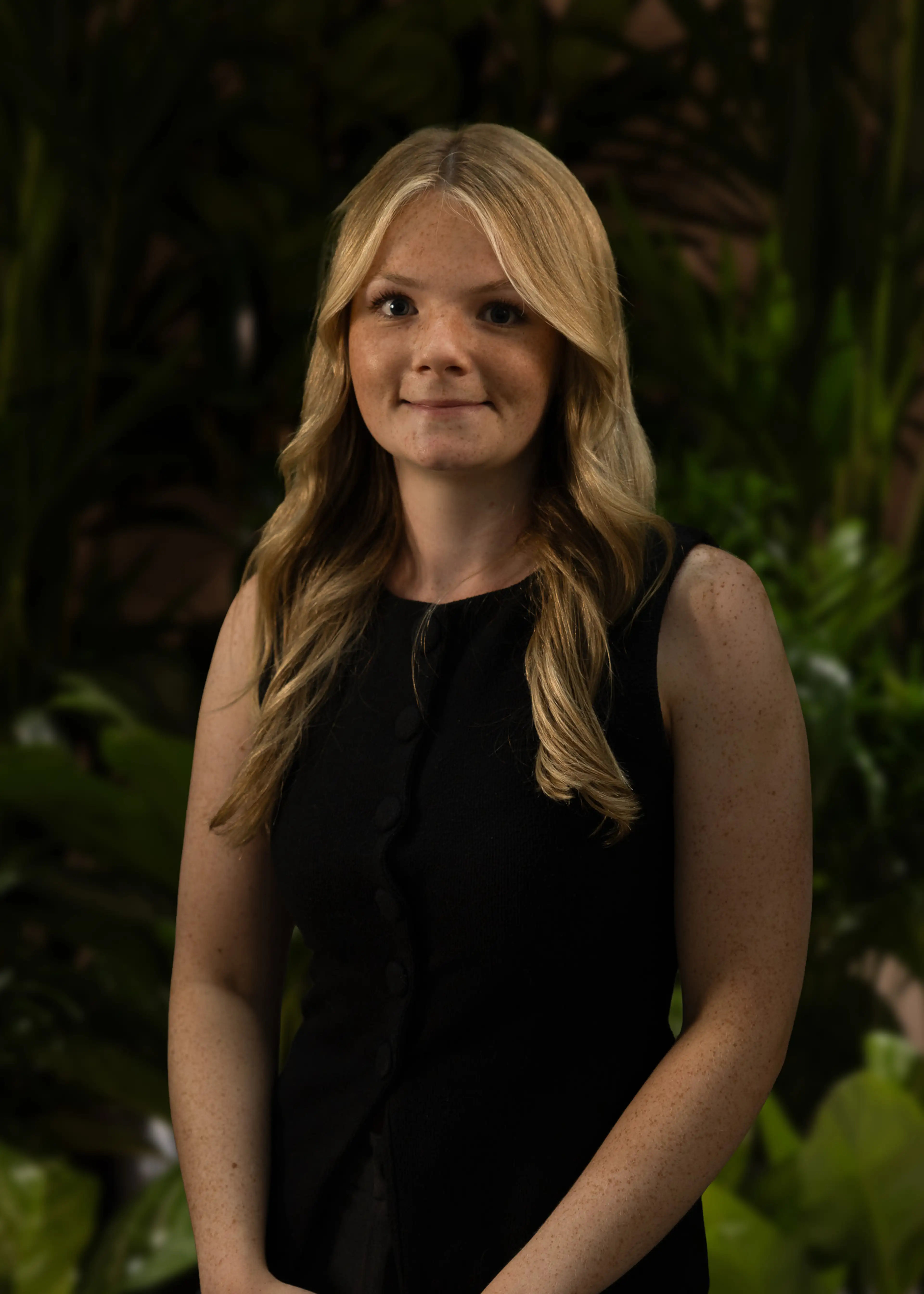 Young woman with long blonde hair in a black sleeveless top standing in front of lush green foliage.