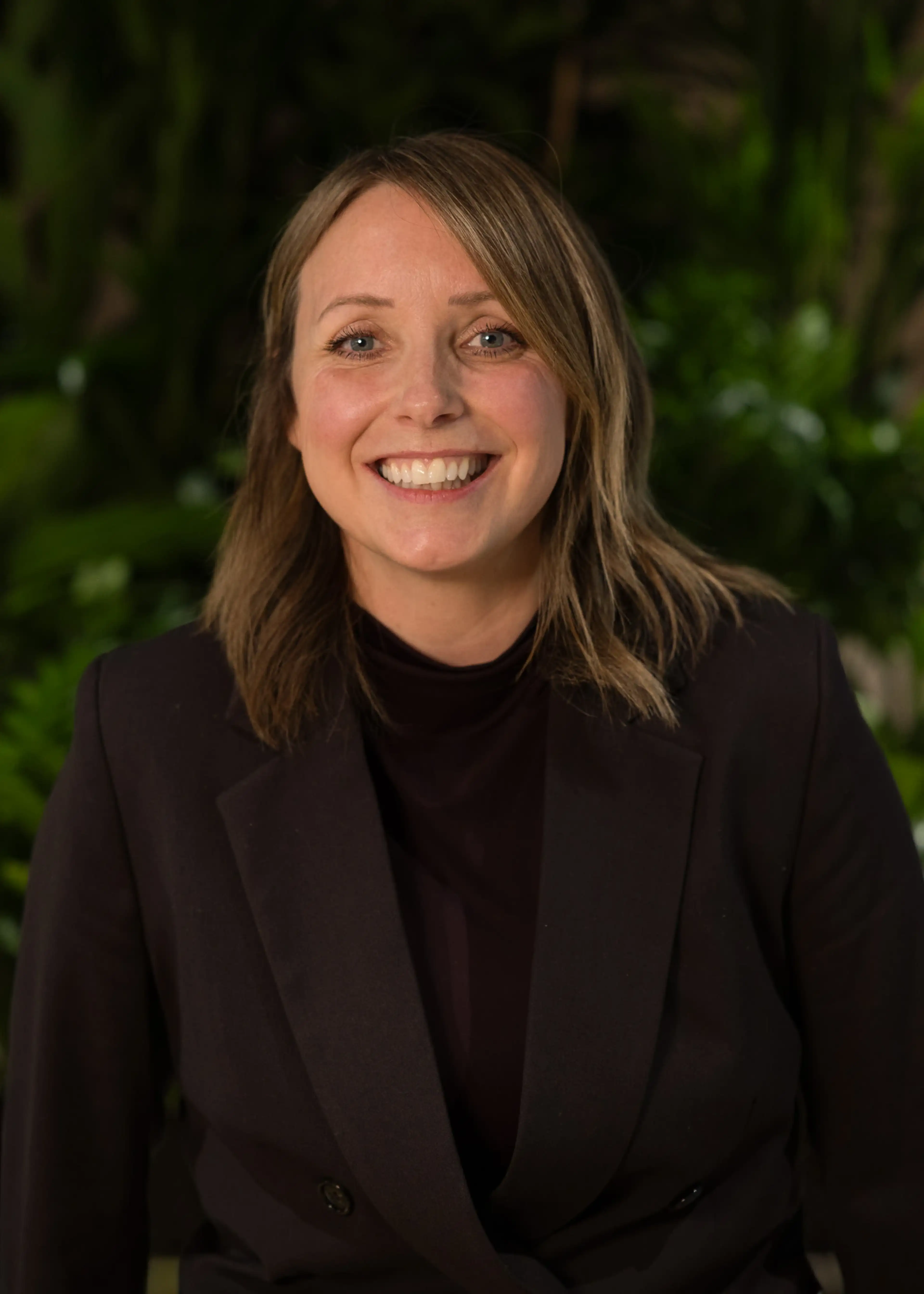 Smiling person with shoulder-length hair in a dark blazer, standing in front of a leafy background.
