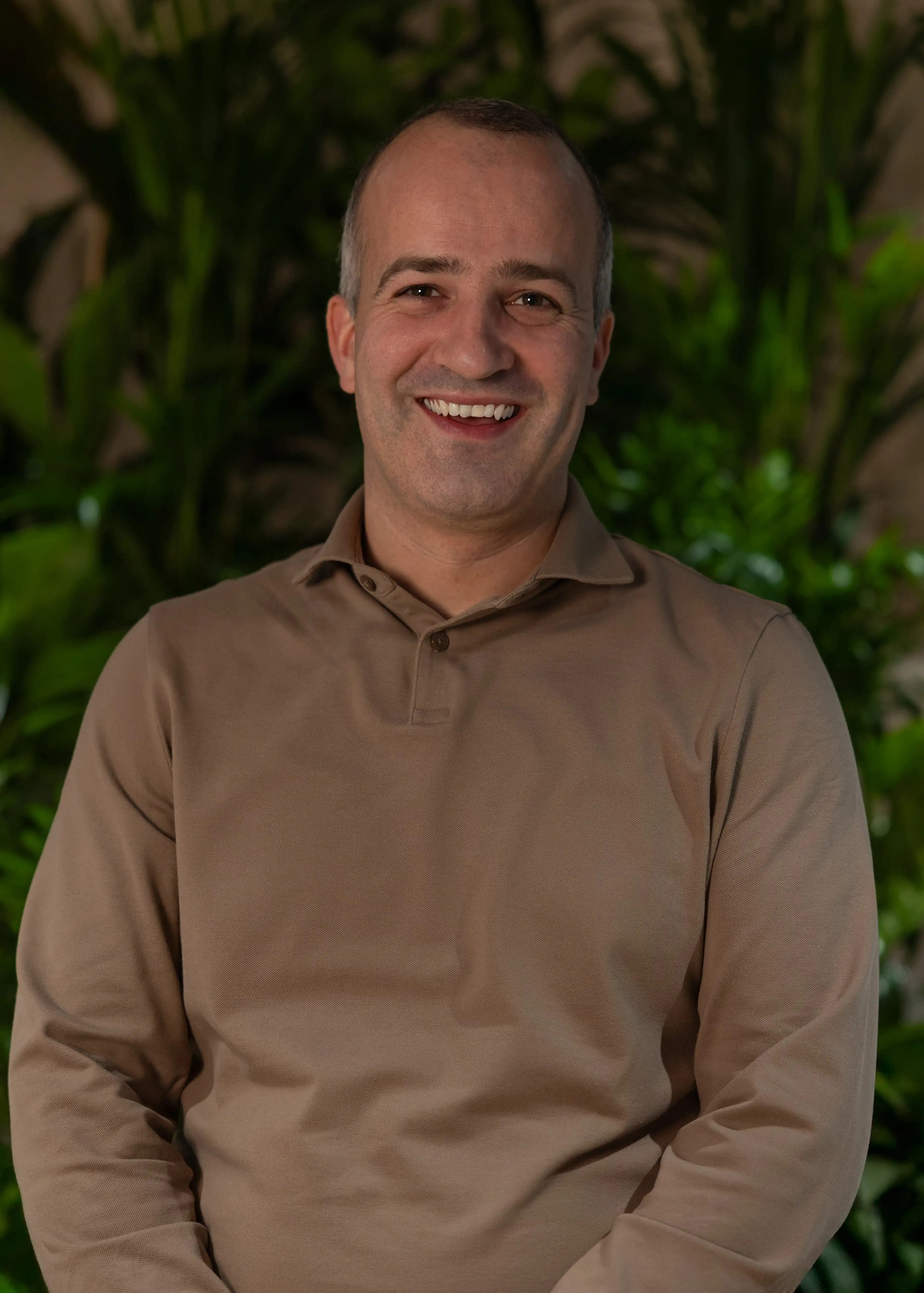 Smiling person in a beige shirt, standing in front of green plants.