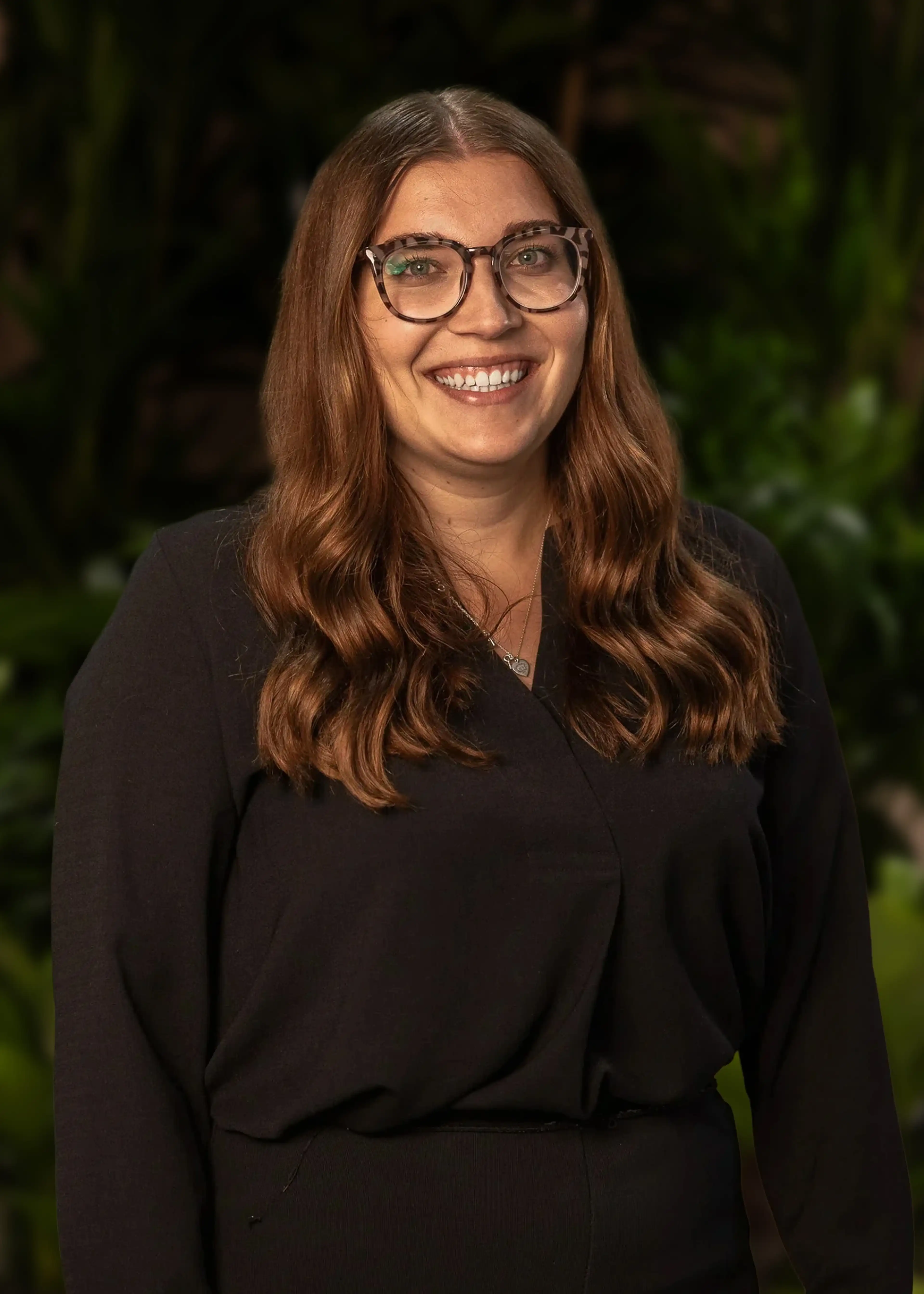 A woman with long brown hair and glasses, wearing a black outfit, smiles while standing in front of a leafy green background.