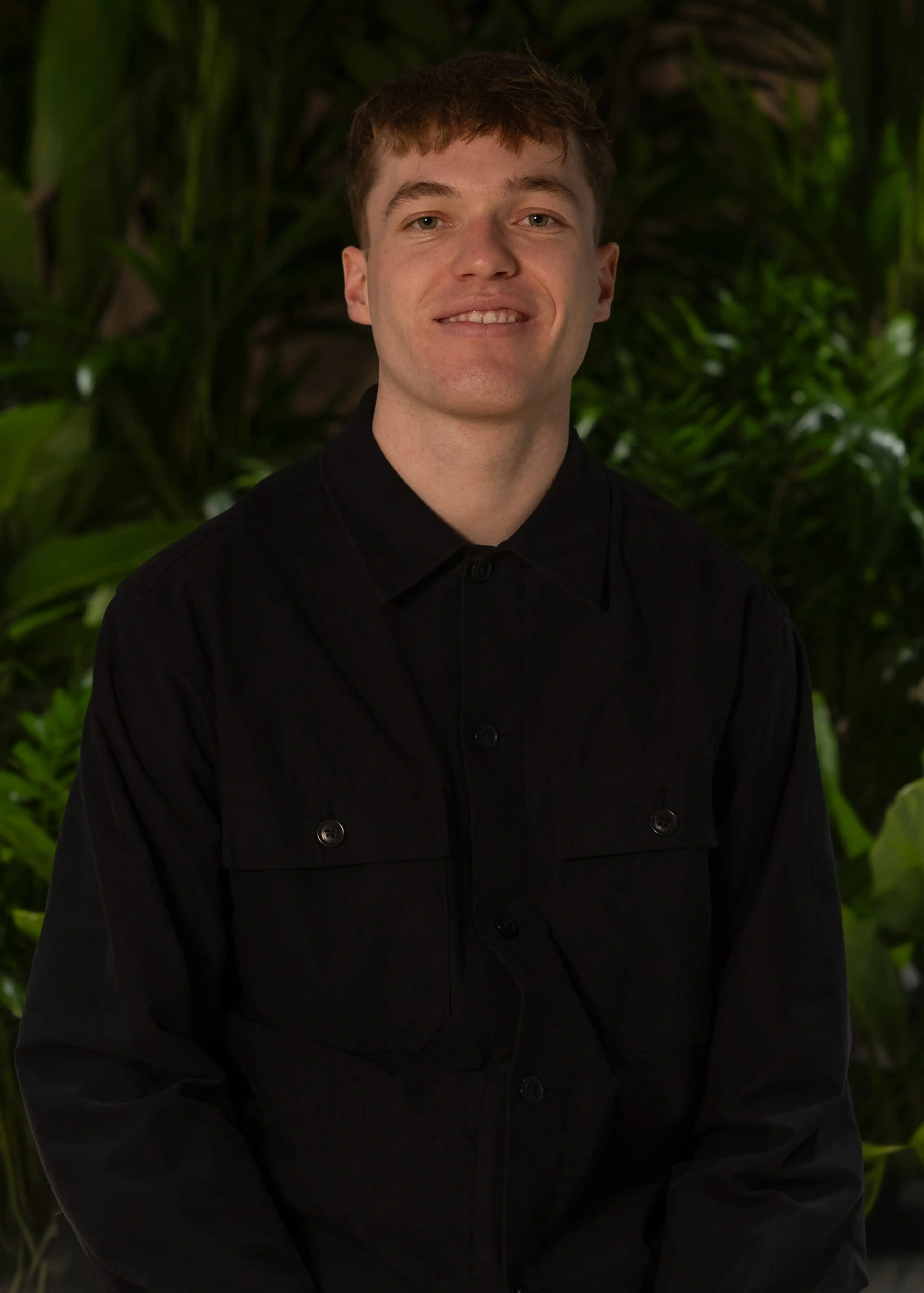 Person in a black button-up shirt smiling, standing in front of lush green foliage.