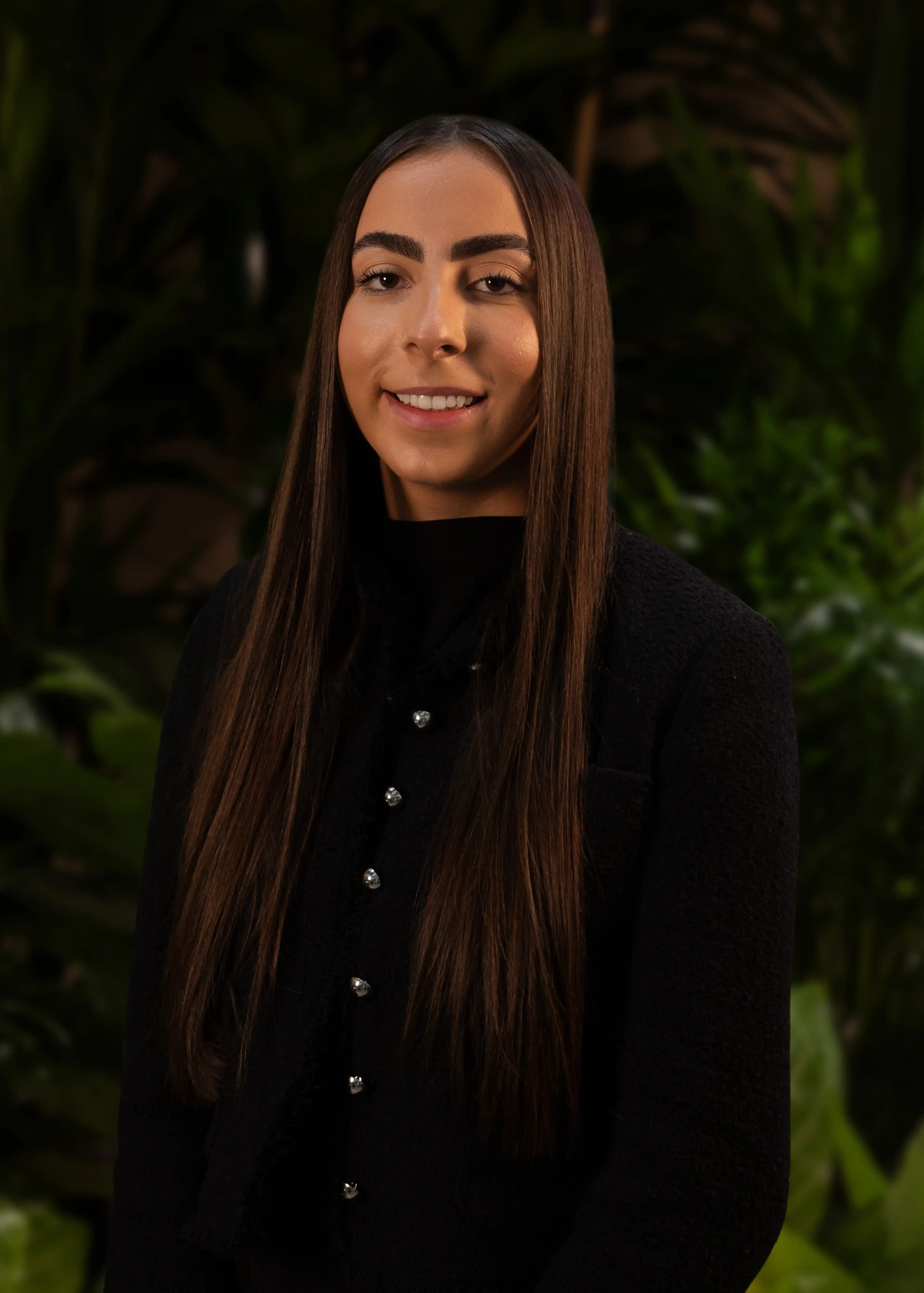 Smiling person with long brown hair in a black jacket, standing in front of a leafy green background.