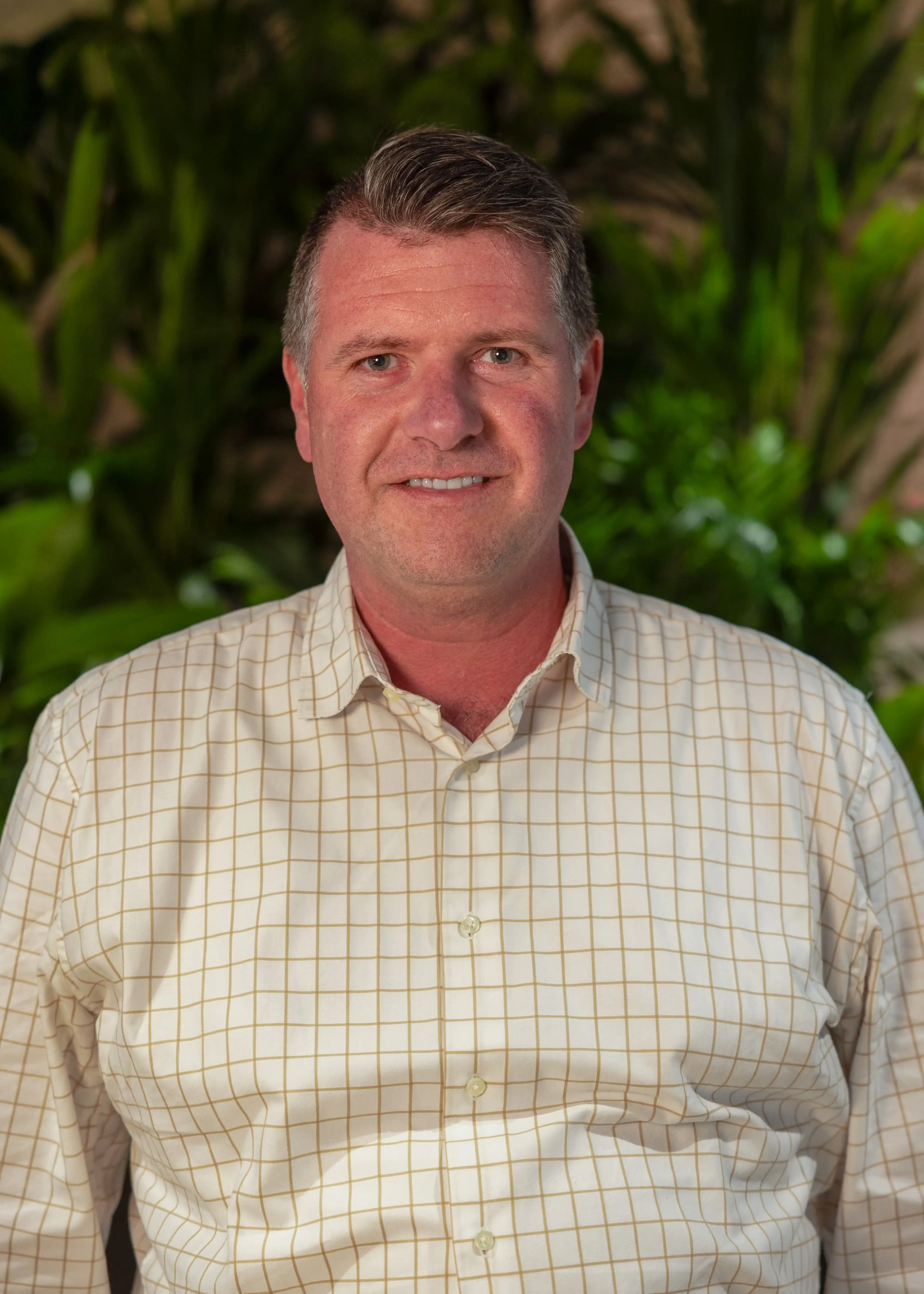 A man in a checkered shirt smiling, standing in front of a background with green plants.