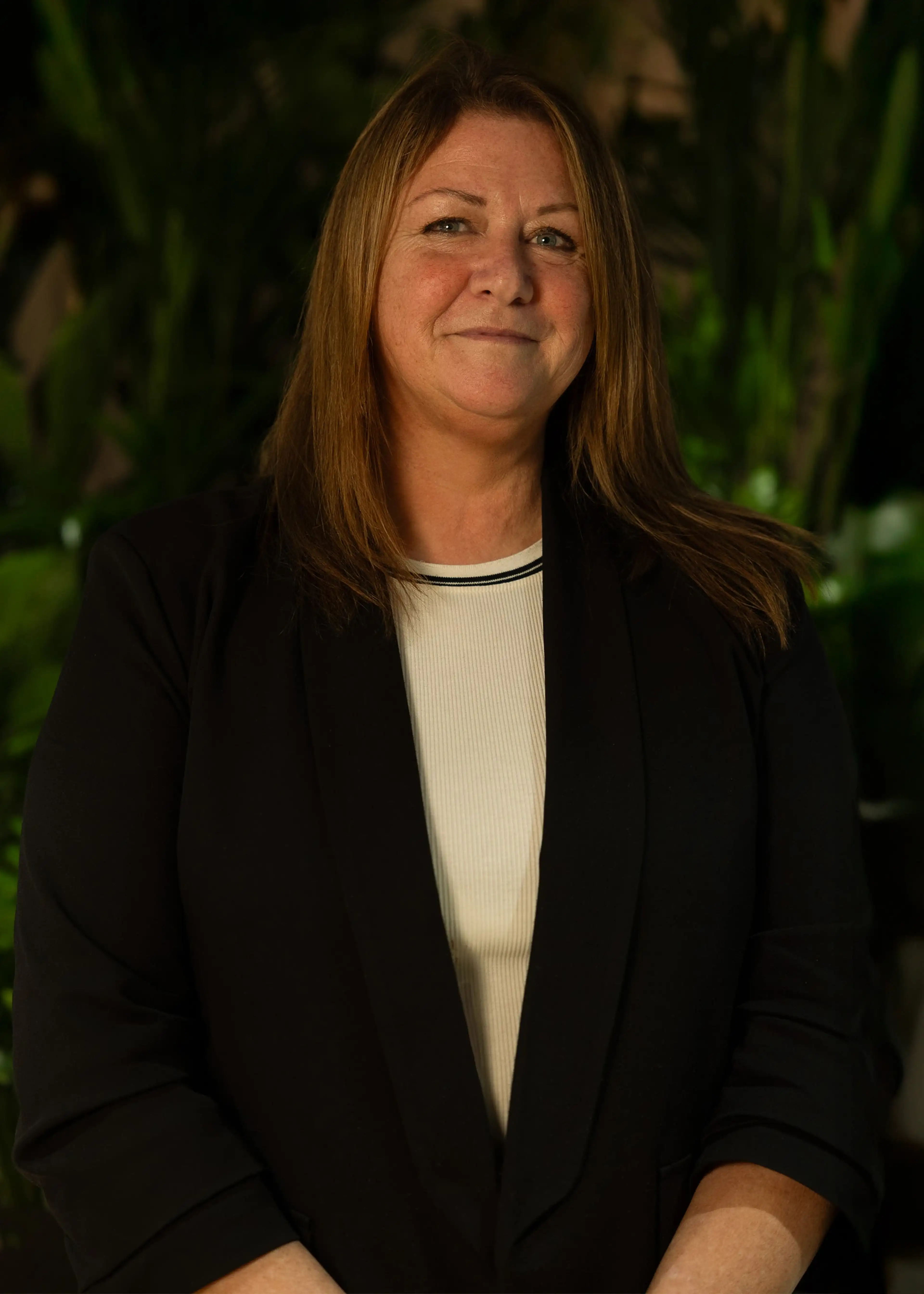 Smiling woman with long brown hair wearing a black blazer over a white top, standing in front of a dark green leafy background.