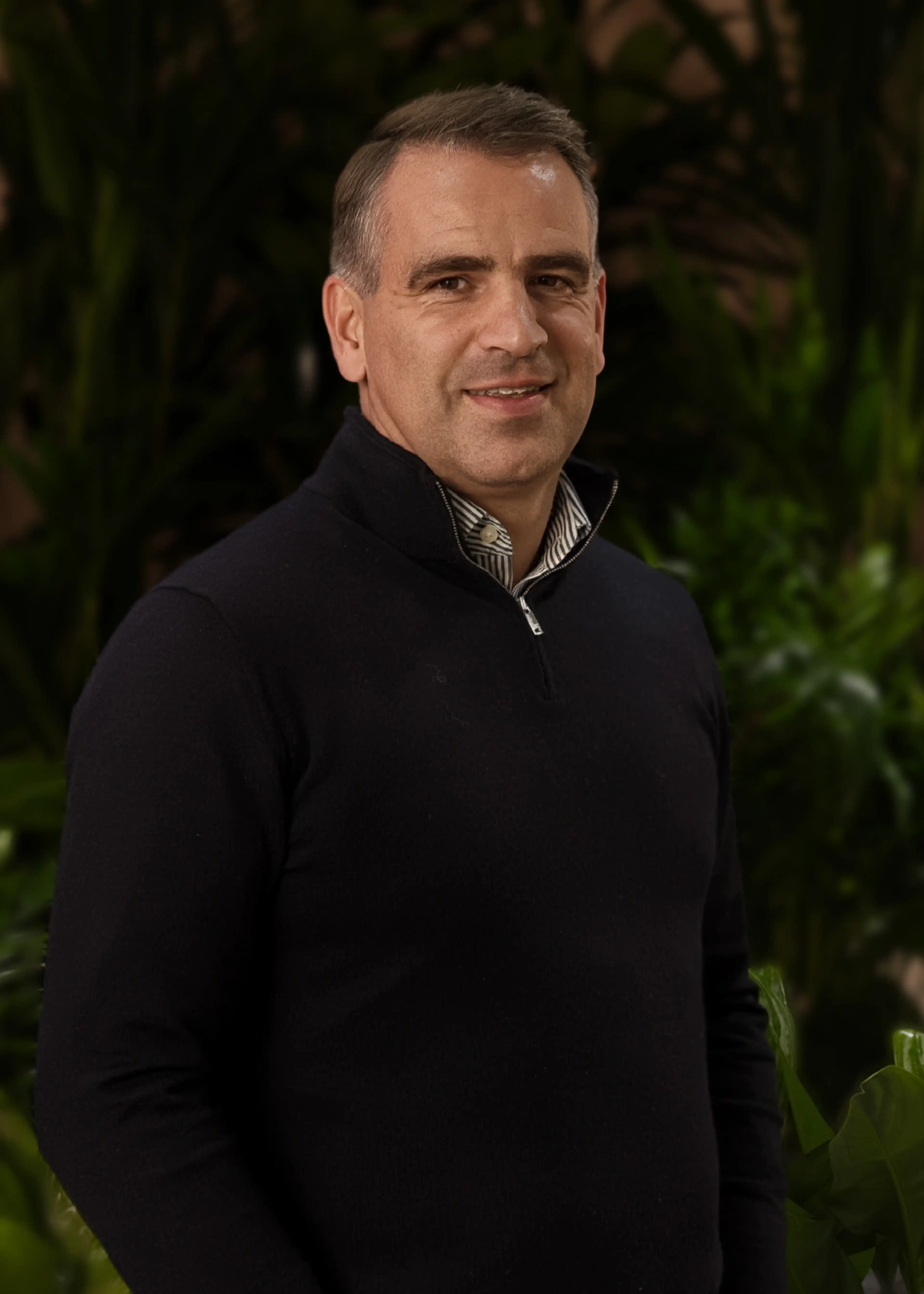 A man wearing a dark sweater stands smiling in front of lush green foliage.