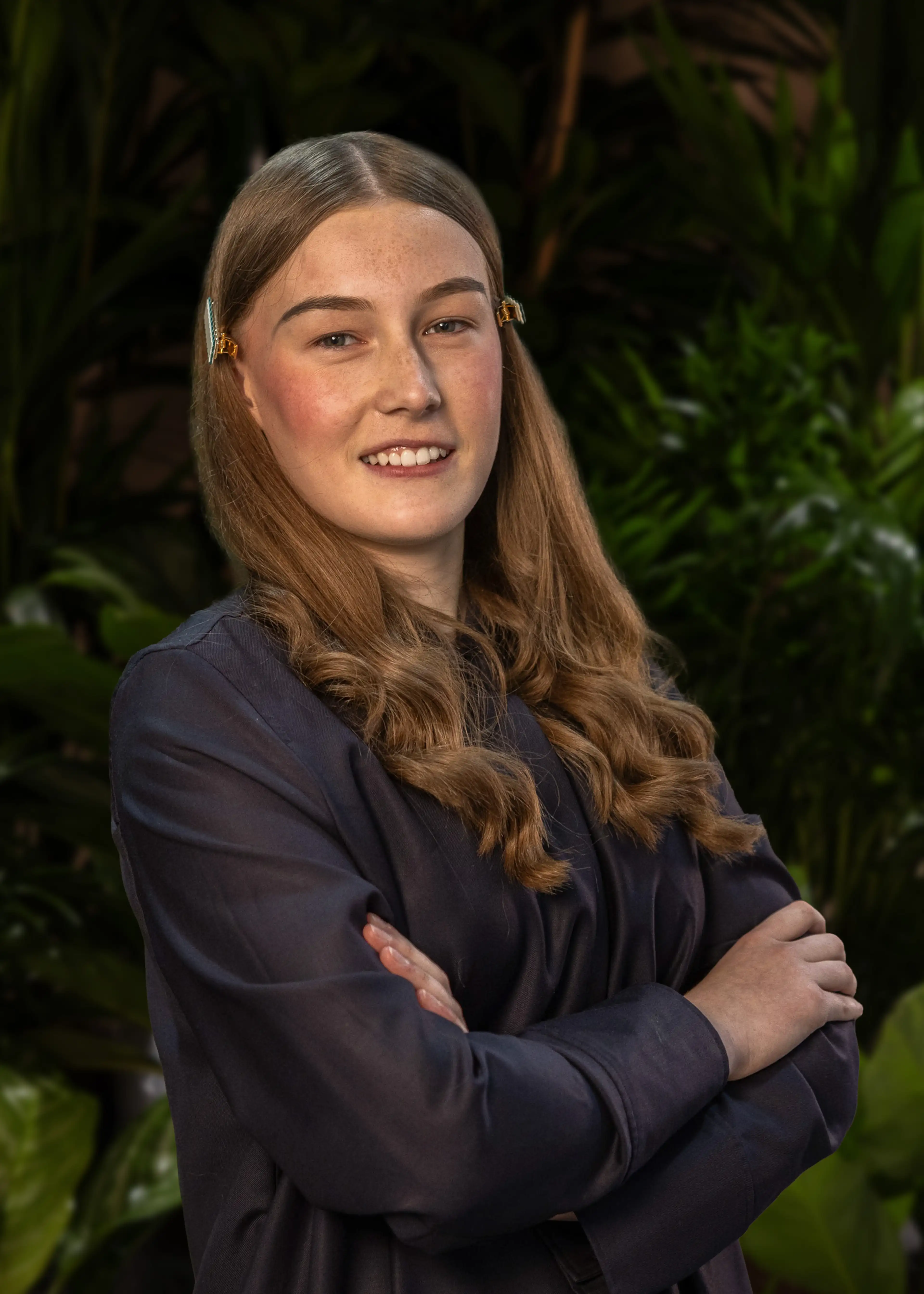 Young woman with long hair, wearing a dark shirt, stands confidently with arms crossed, surrounded by lush green plants.