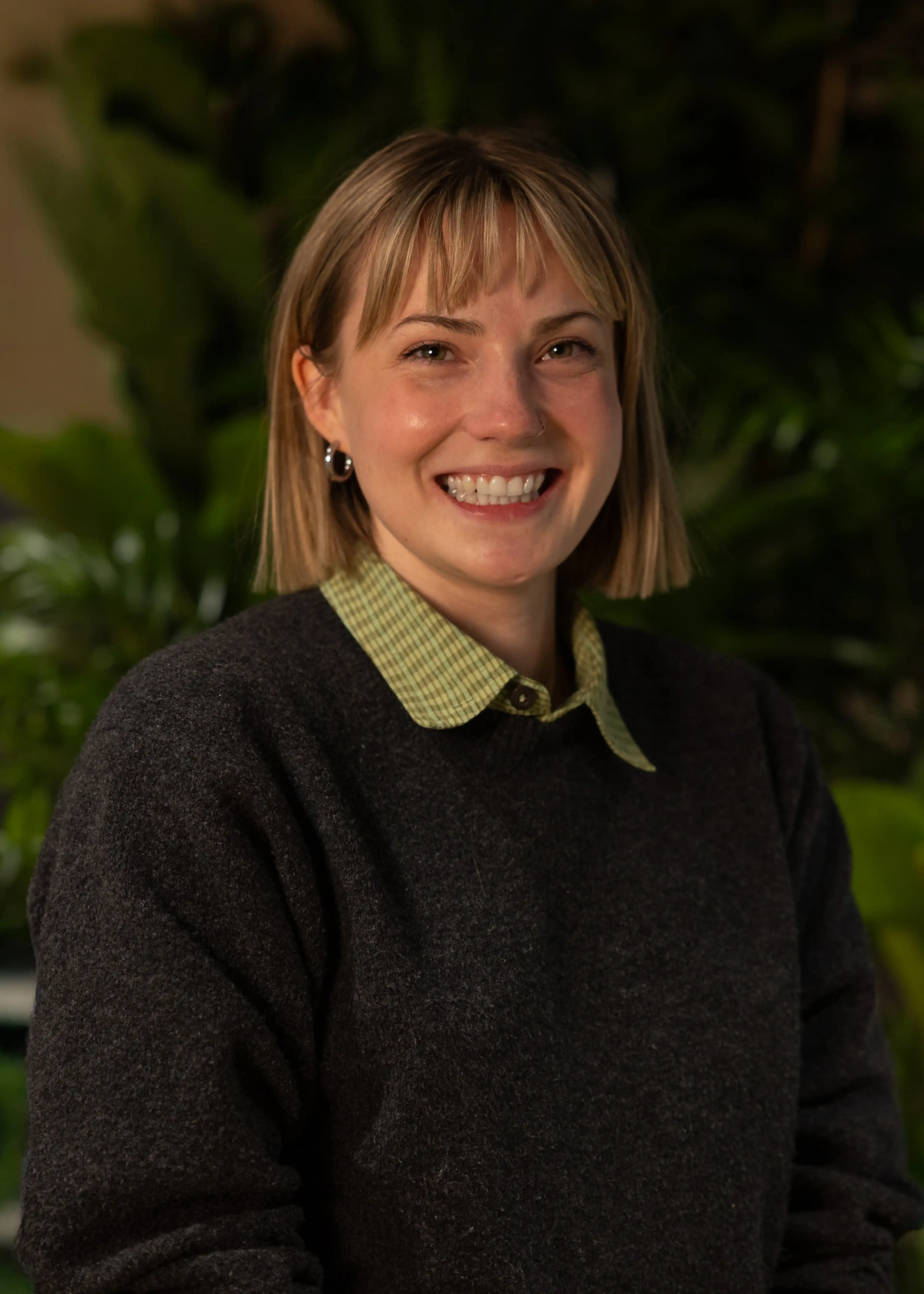 Smiling person with short blond hair, wearing a dark sweater over a green checked shirt, standing in front of leafy plants.