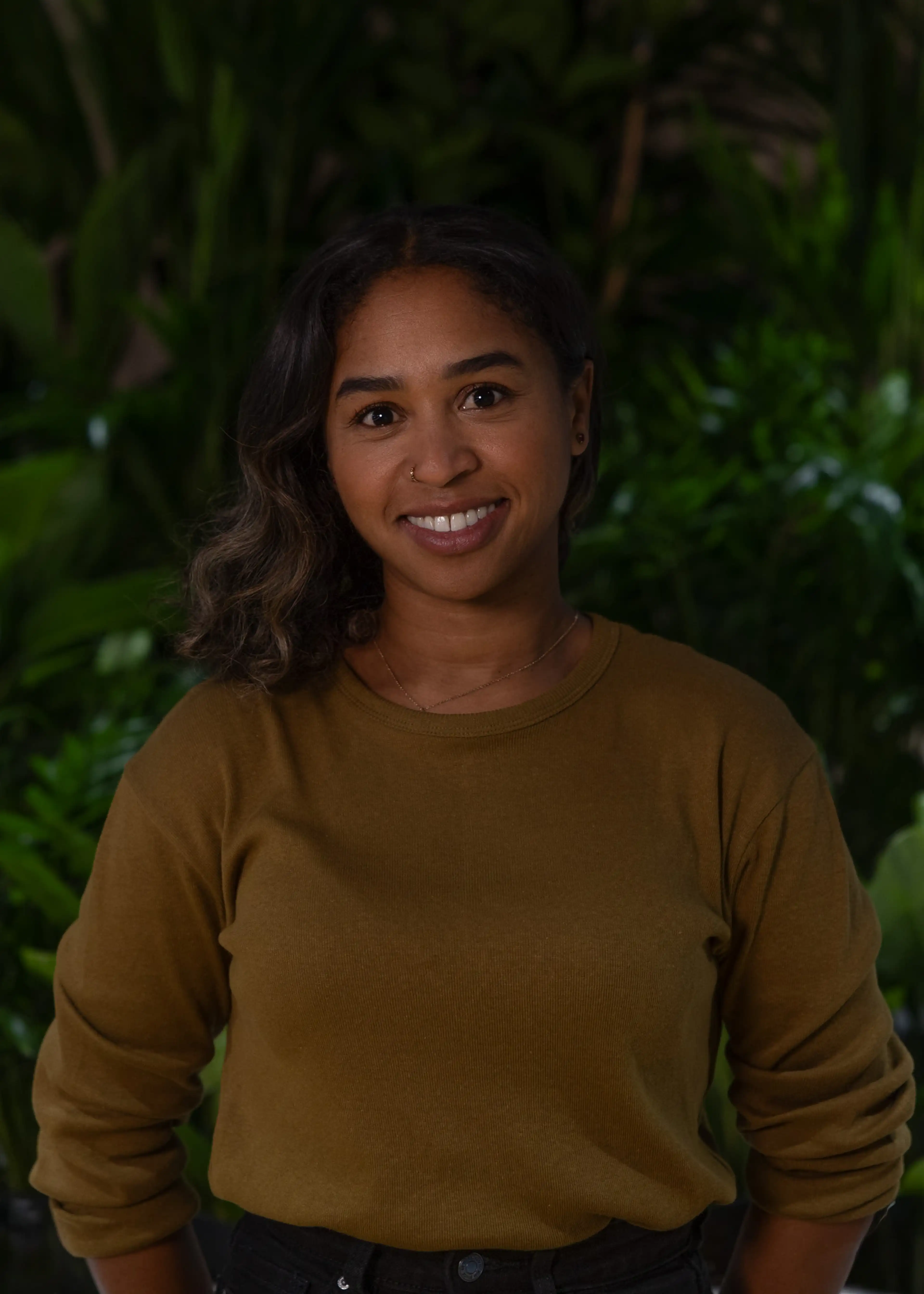 A woman in a brown sweater stands smiling with her arms at her sides against a lush green plant background.
