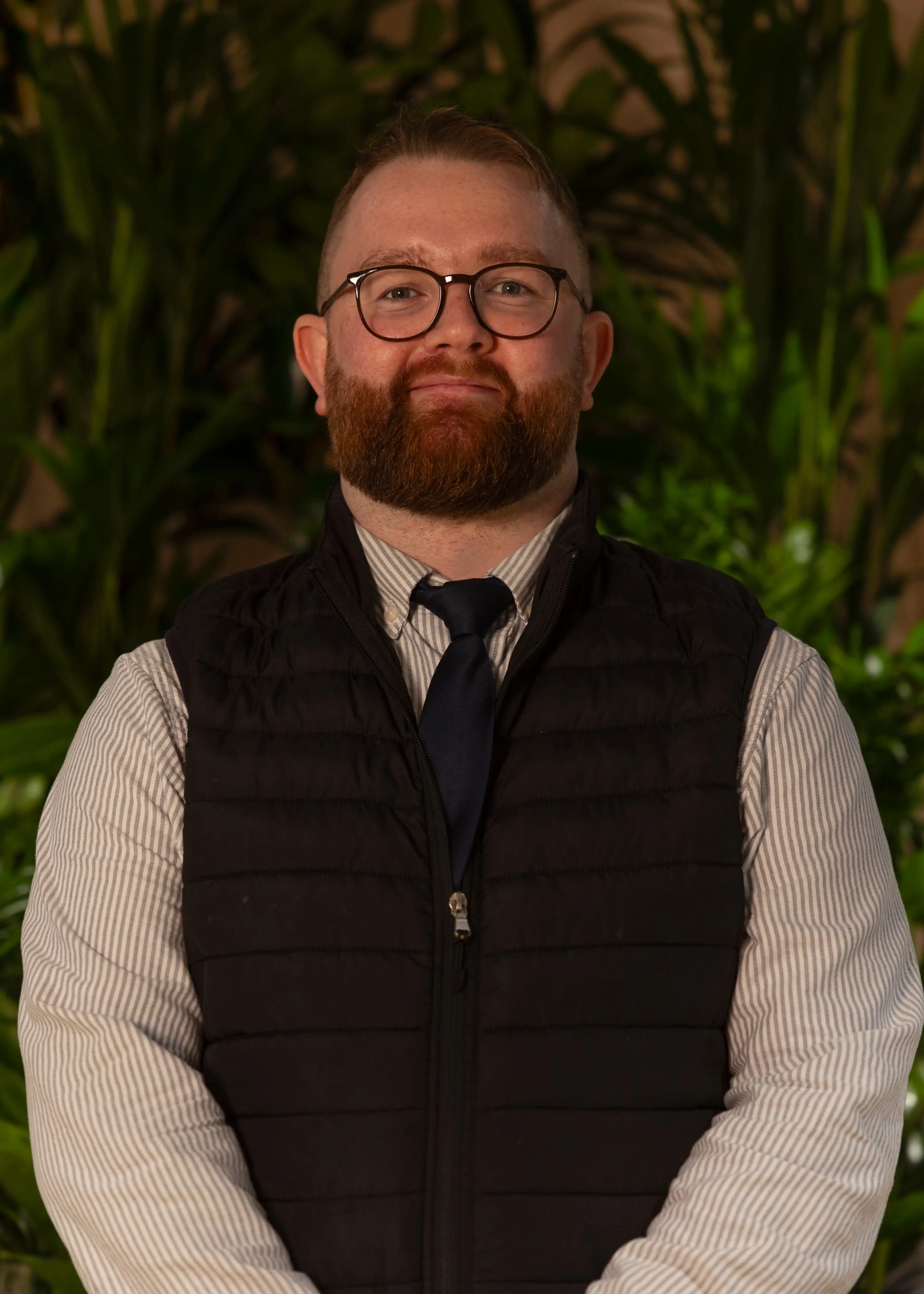 Man with glasses and beard, wearing a striped shirt, tie, and black vest, stands in front of lush green plants, smiling.