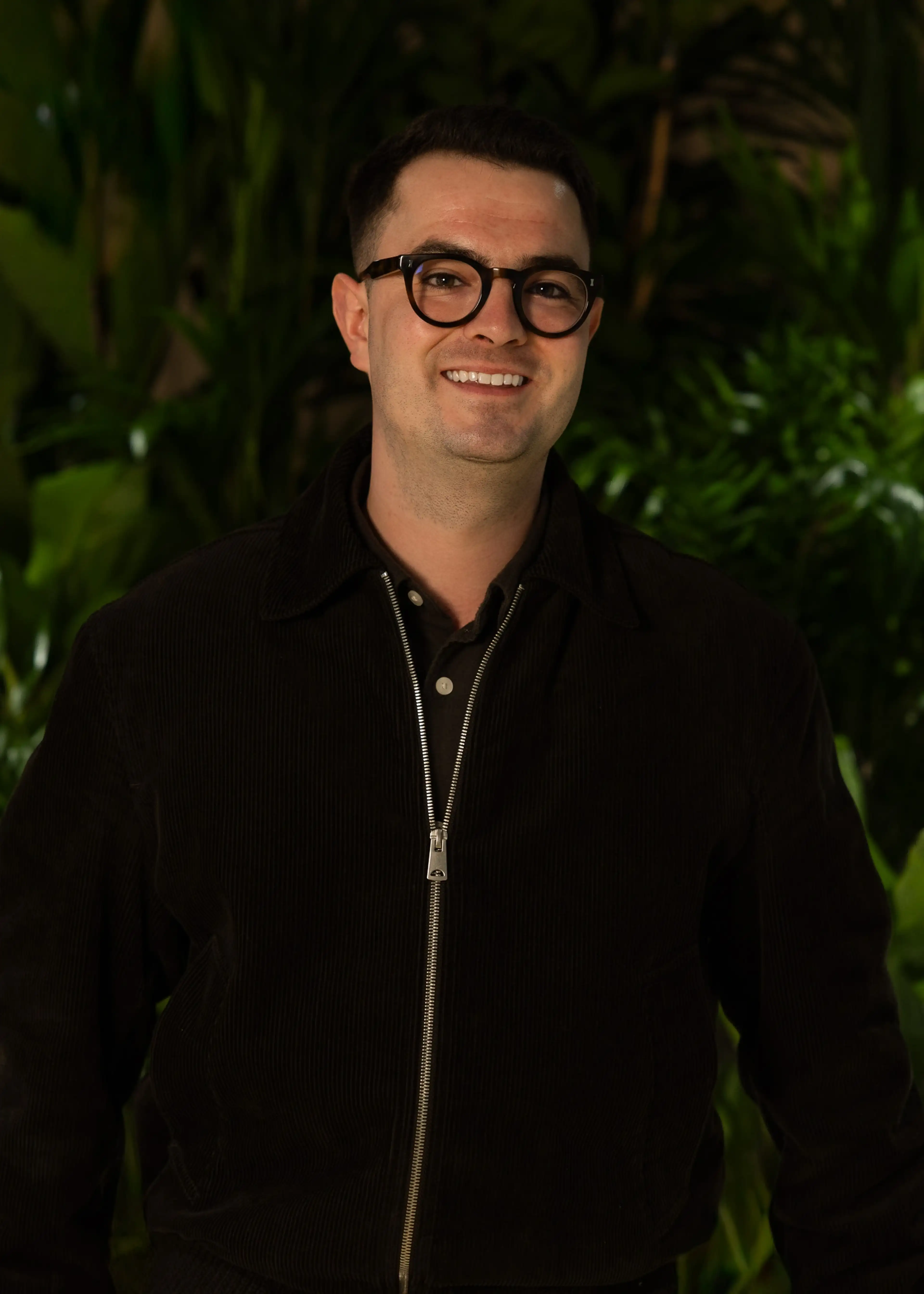 Person wearing glasses and a dark jacket smiles in front of lush green foliage.