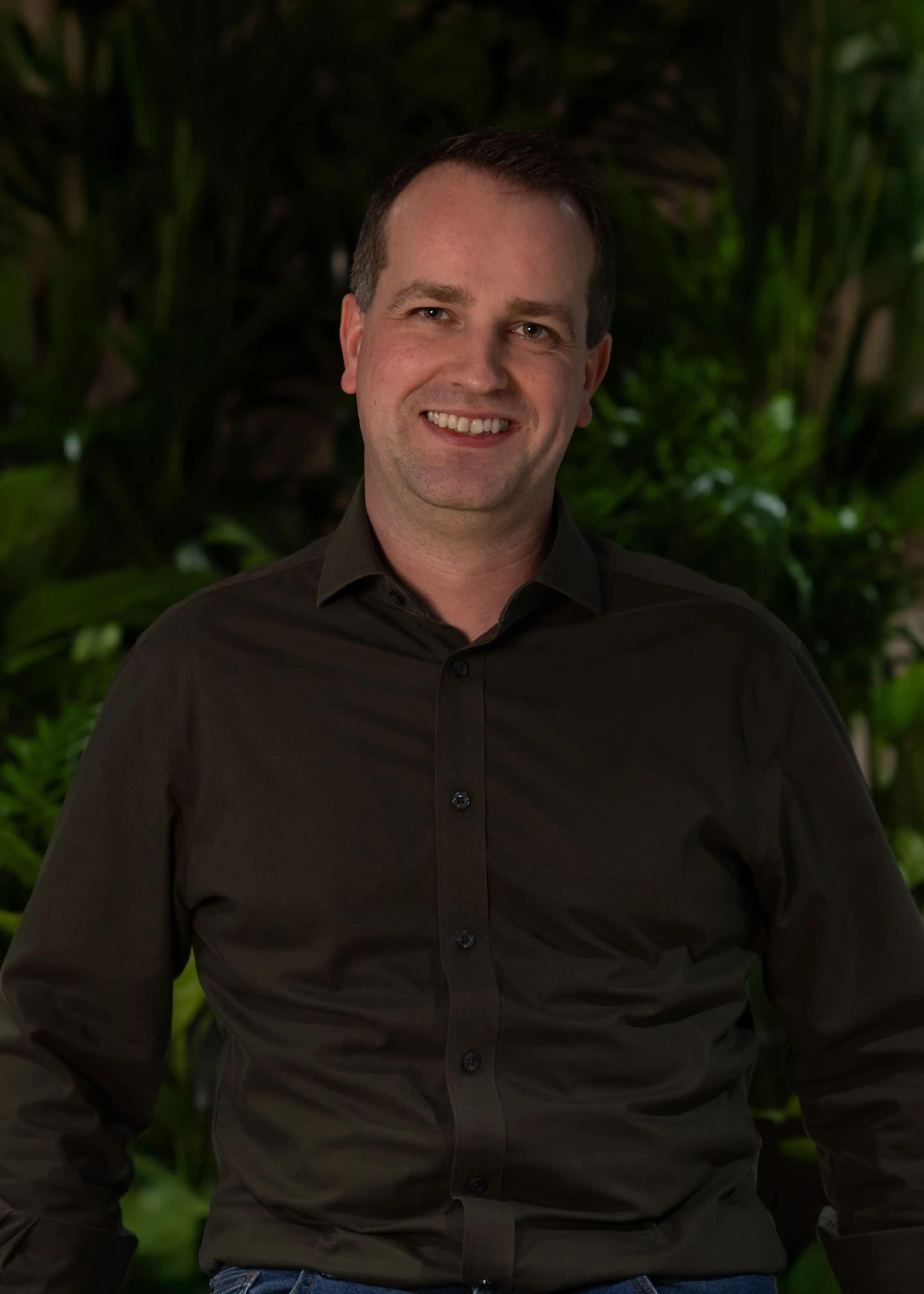 Smiling man in a dark shirt, standing against a backdrop of lush green plants.