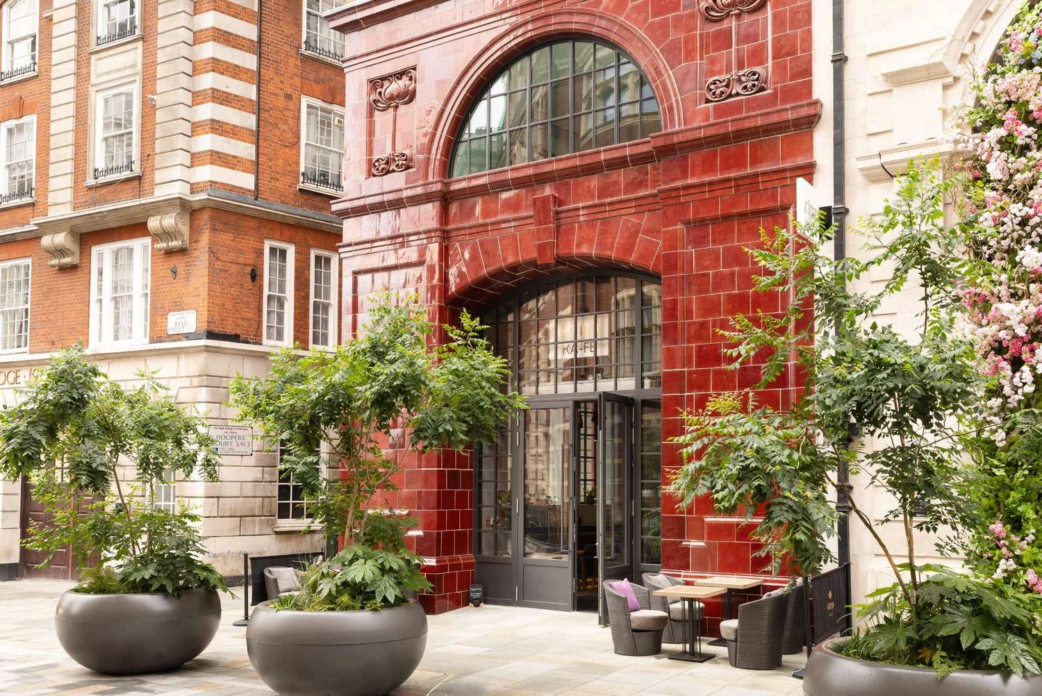 Entrance of a red-tiled building with large windows, outdoor seating, and potted plants on a sidewalk, surrounded by other buildings.