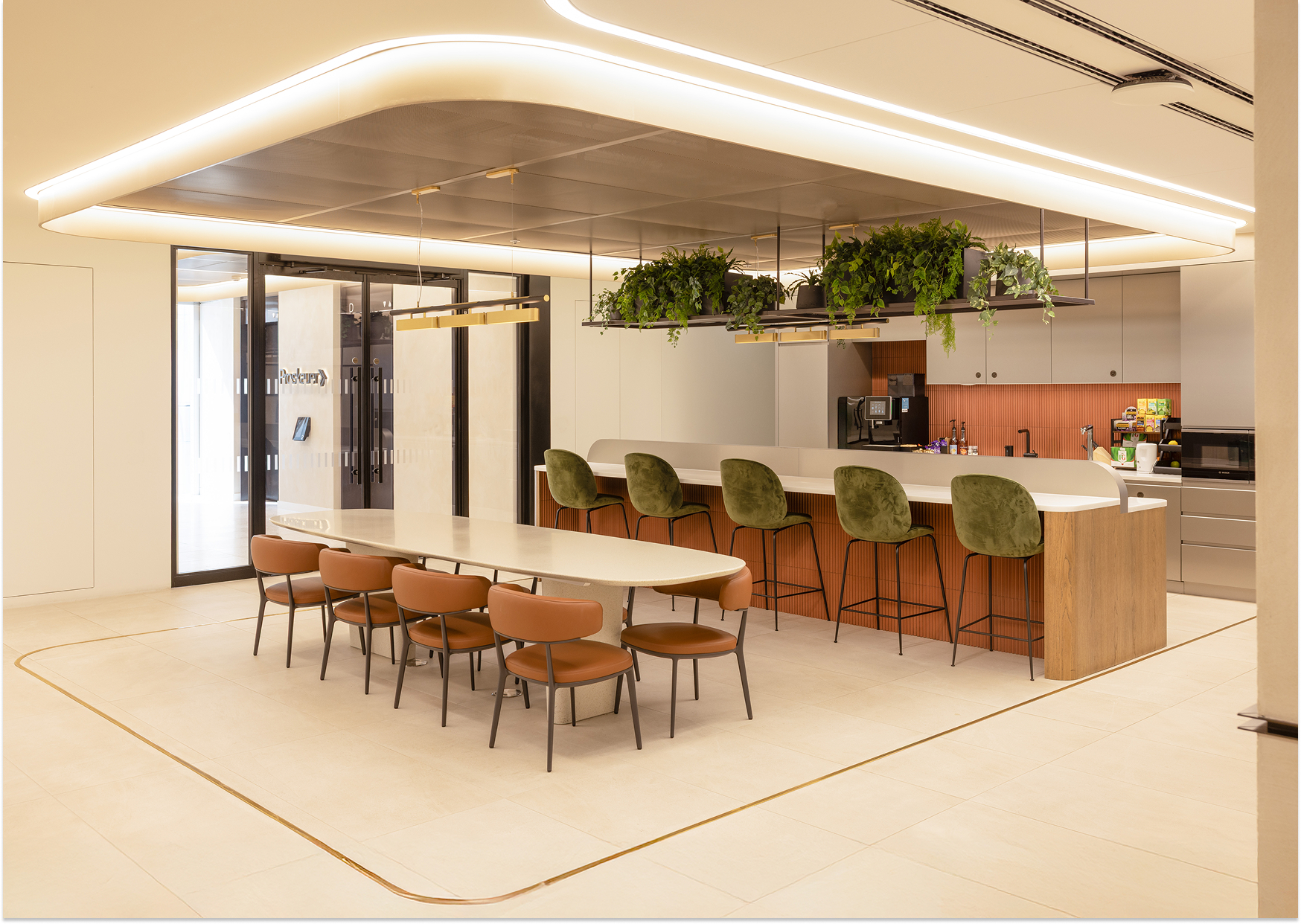 Modern office kitchen with a long table, brown chairs, a bar with green stools, hanging plants, and soft overhead lighting.