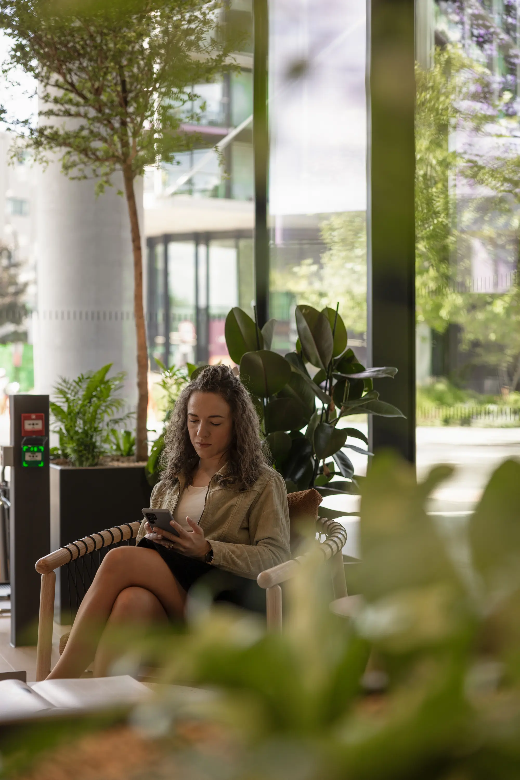 A woman sits on a chair in a modern, plant-filled room, looking at her phone. Large windows reveal a blurred view of the outdoors.