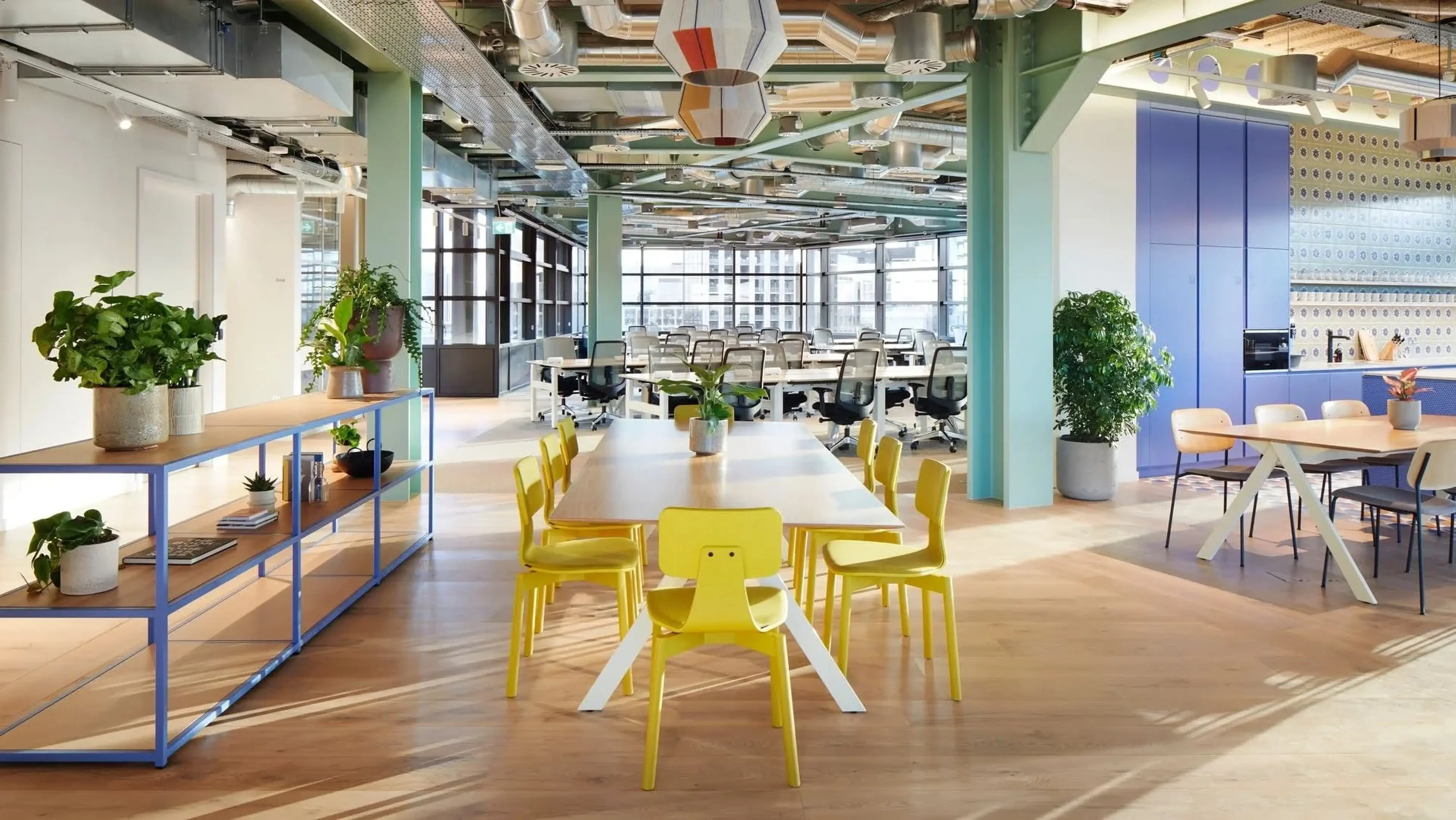 Modern office space with yellow chairs around a wooden table, potted plants, exposed ceiling, and an adjacent kitchen area with blue cabinets.