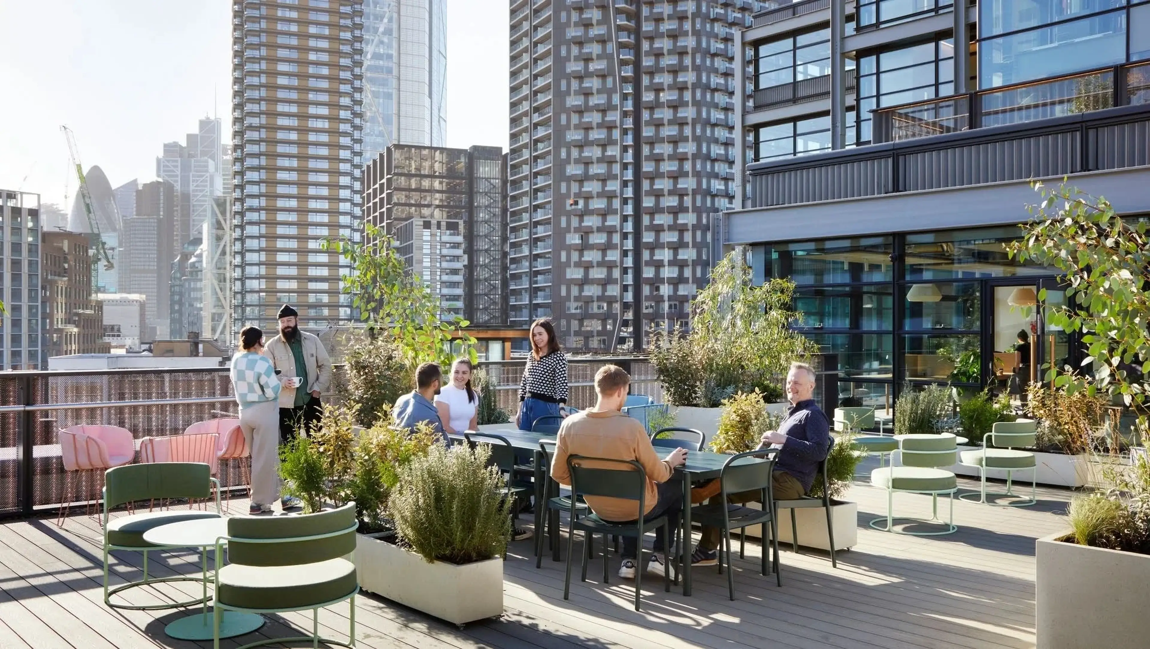 People socializing on a rooftop terrace with city skyscrapers in the background. The area has tables, chairs, and greenery.