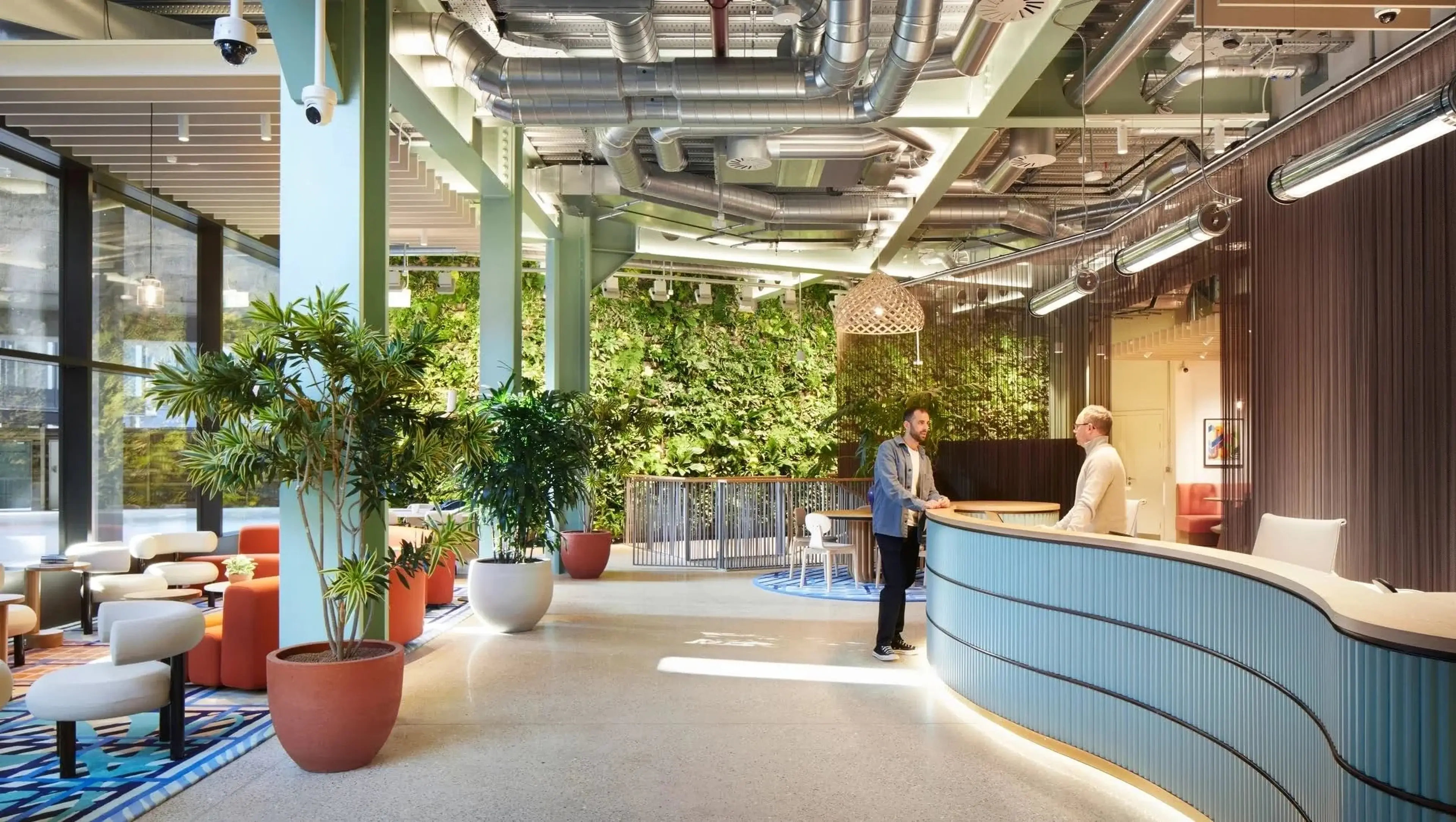 Modern office lobby with green wall, large plants, and a curved front desk, where two people are conversing. Ductwork is visible on the ceiling.