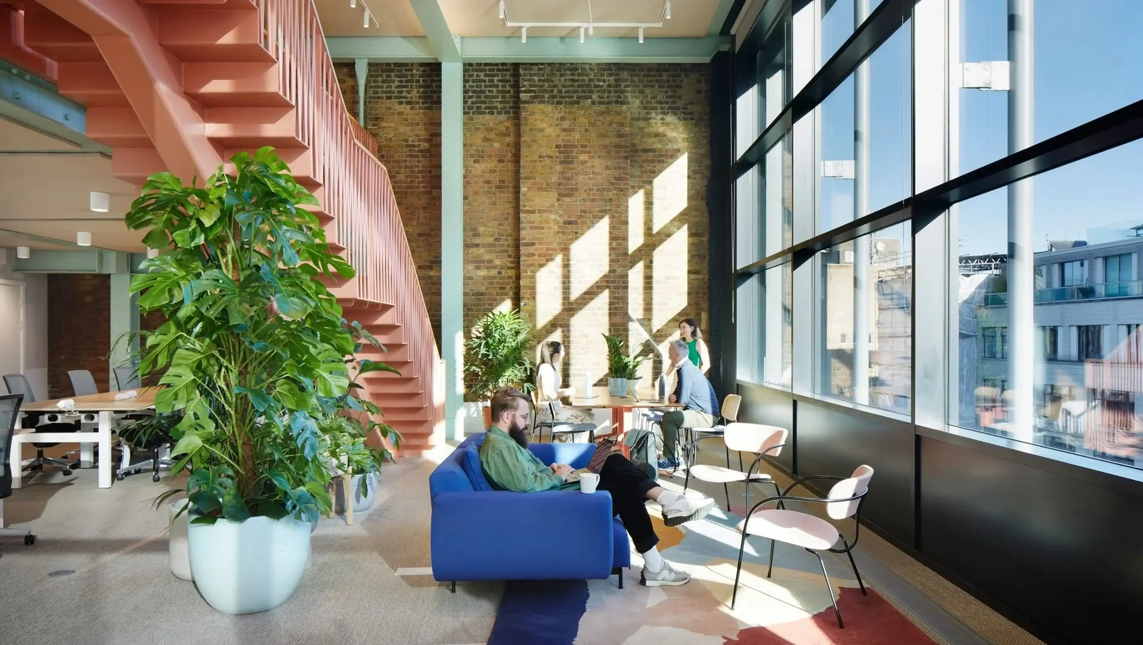 Modern office space with large windows, a pink staircase, green plants, and people working and relaxing on a blue sofa and around a table.