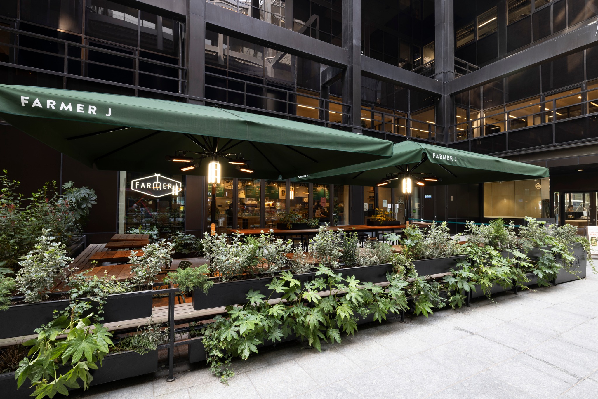 Outdoor seating area of Farmer J restaurant with green umbrellas, benches, and potted plants in an urban setting.