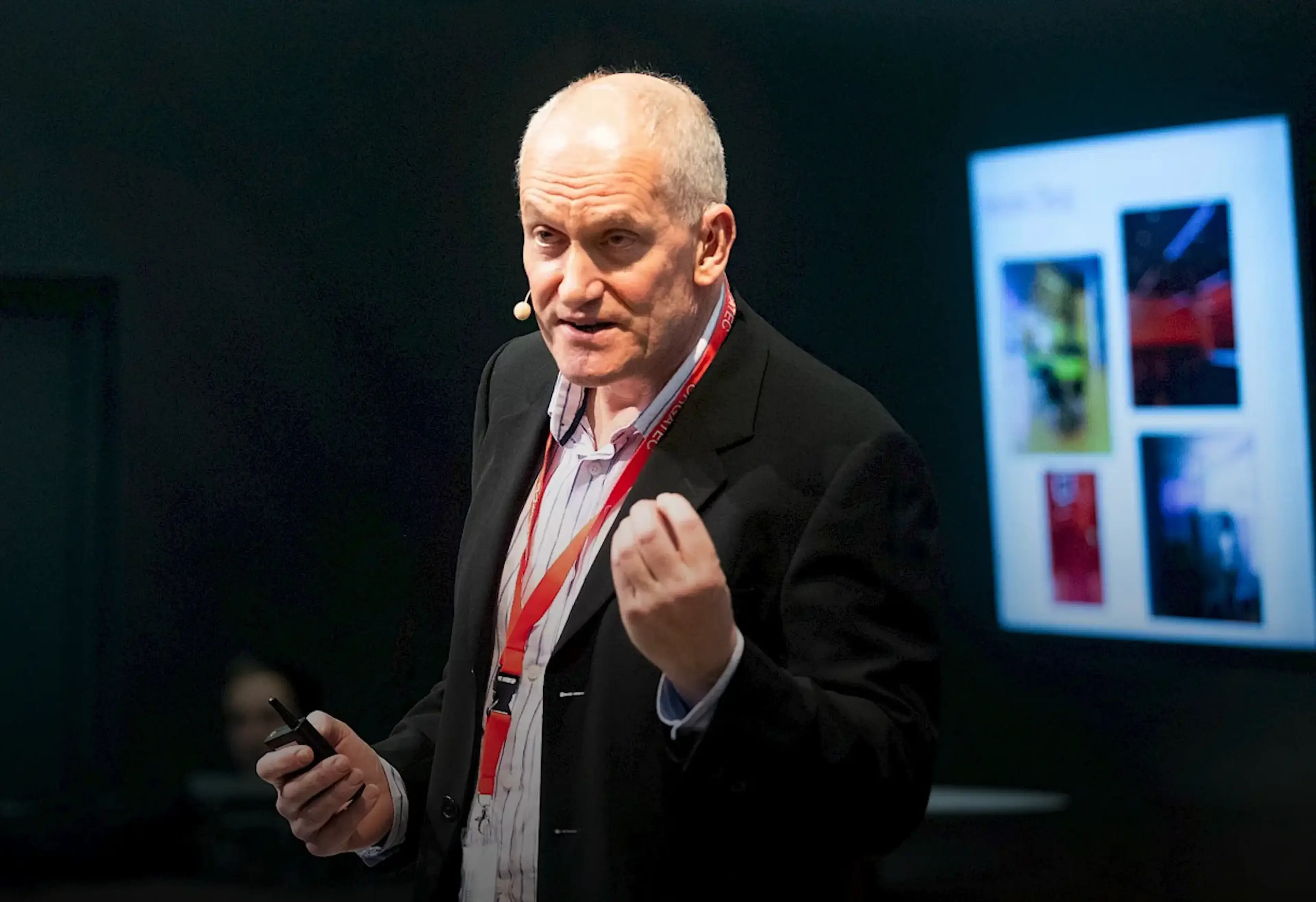 A person with a headset speaks passionately on stage. A screen with images is visible in the background.