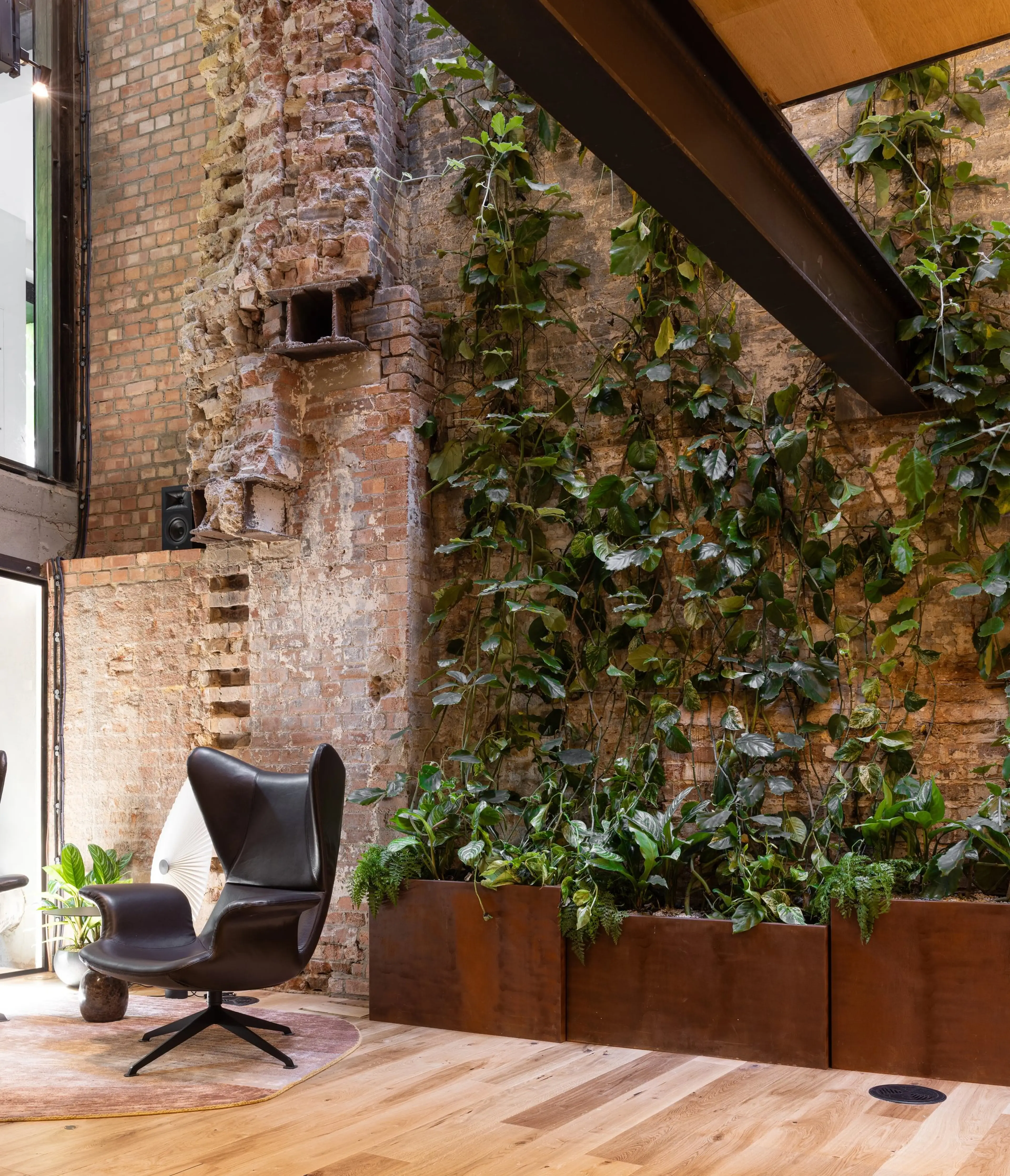 Modern interior with exposed brick wall, vertical garden, and black leather chair on a wooden floor. Natural light from large windows.