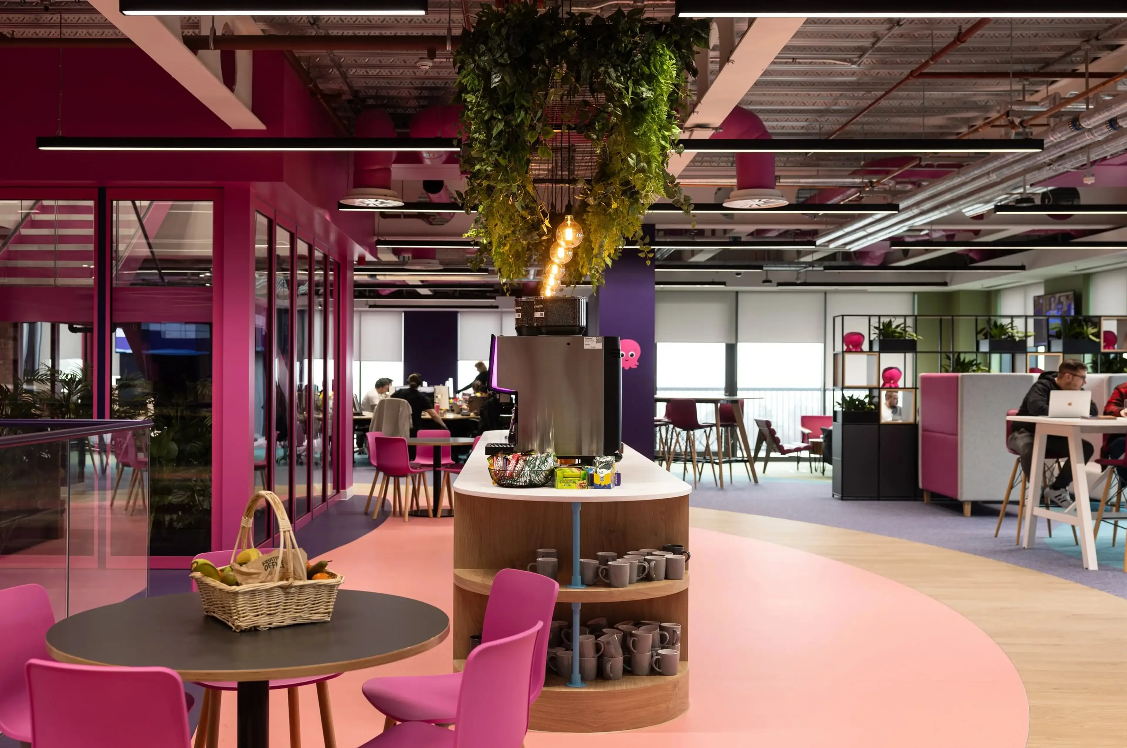 Office coffee bar with vibrant pink and purple decor, hanging plants, round tables, and a snack station. People are working in the background.