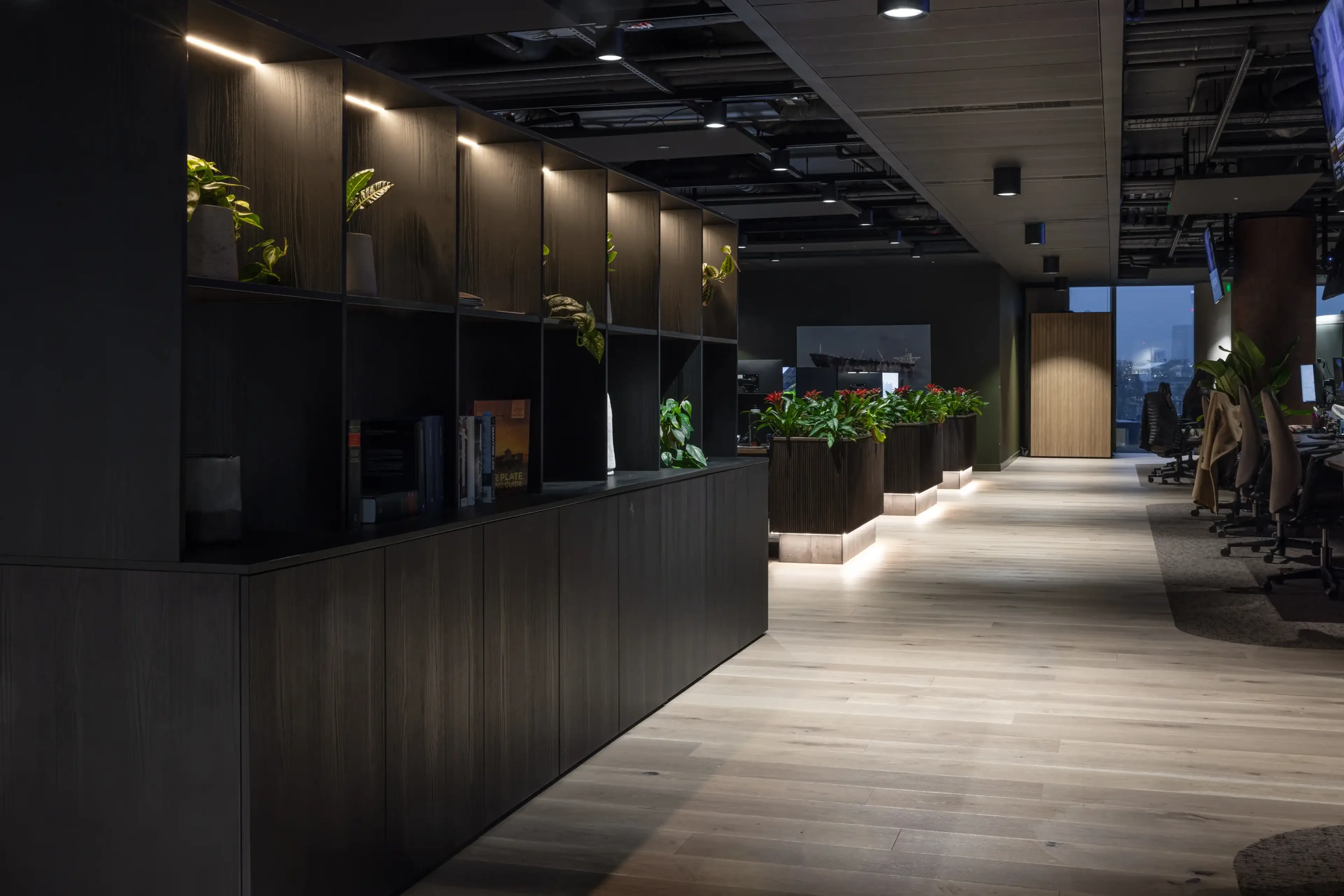 A modern office space with a dark-toned shelving unit featuring built-in lighting, housing small potted plants and books. A row of large floor-standing planters filled with lush greenery and red bromeliads lines the corridor, illuminated by underlighting.