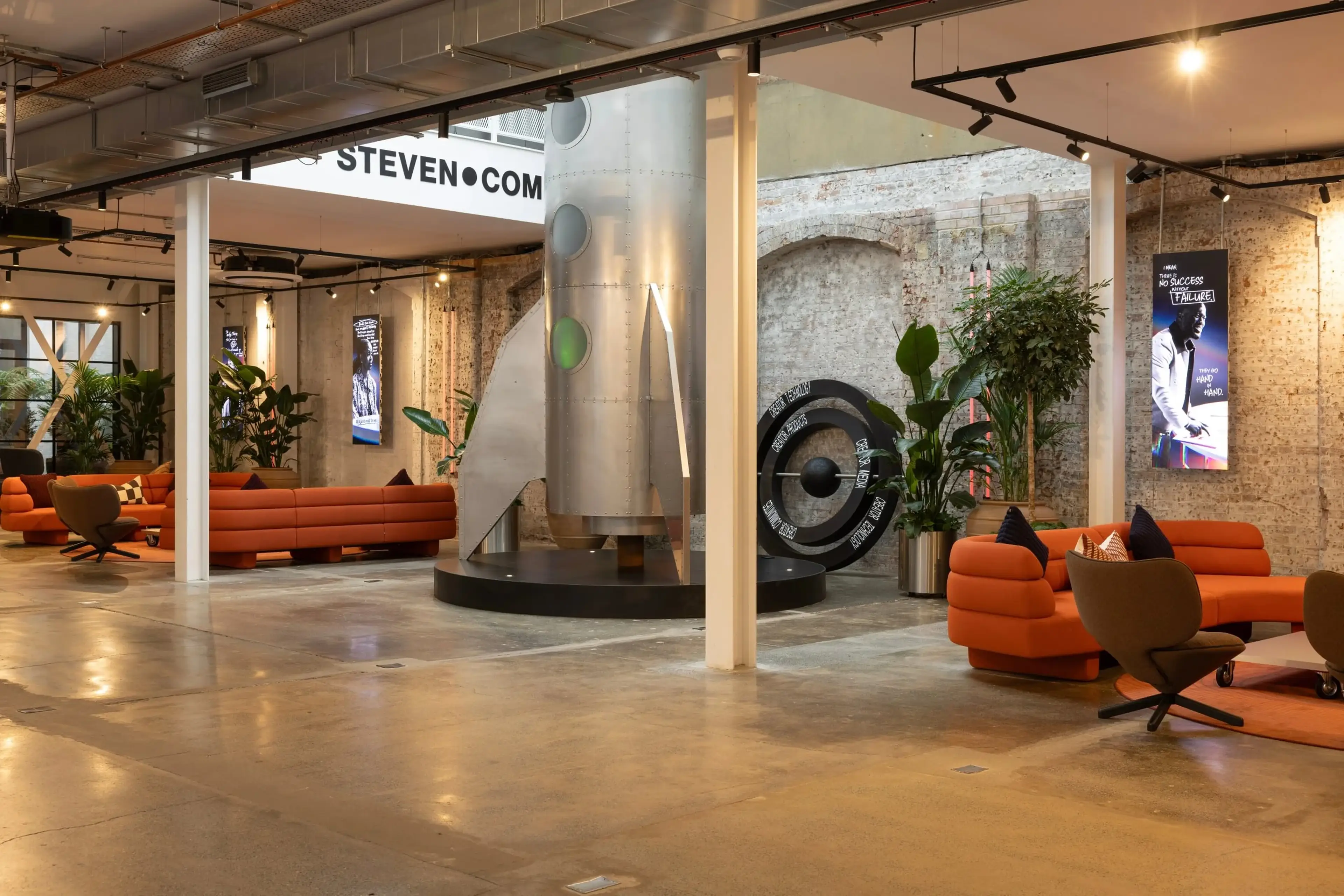 Modern office space with orange sofas, a silver rocket sculpture, greenery, and digital screens on exposed brick walls.